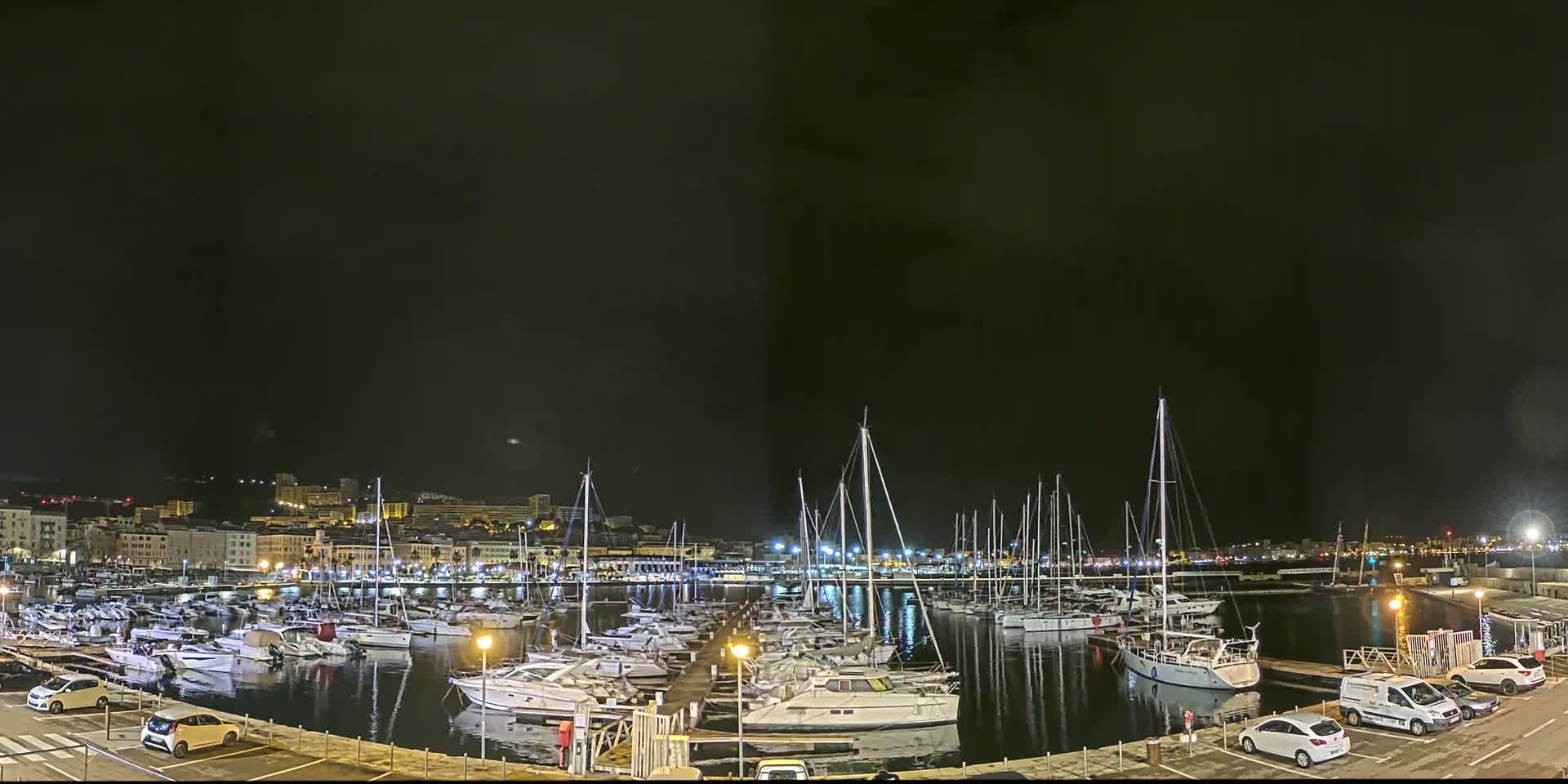 A nighttime view of a marina filled with sailboats and motorboats, docked along a waterfront with a city skyline in the background under a dark sky.