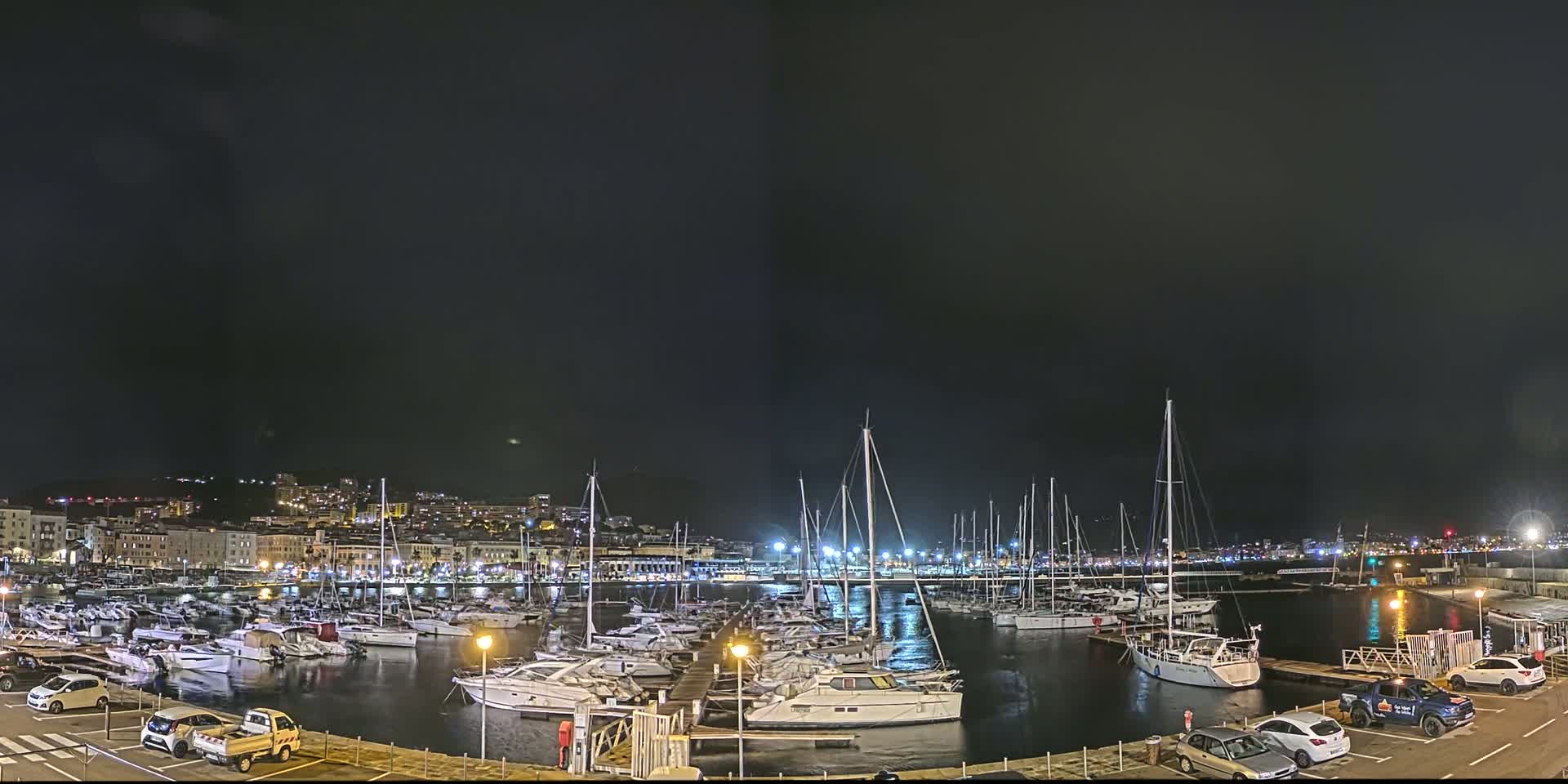 A nighttime view of a marina filled with numerous boats, docked alongside a city's illuminated waterfront under a dark, clear sky.
