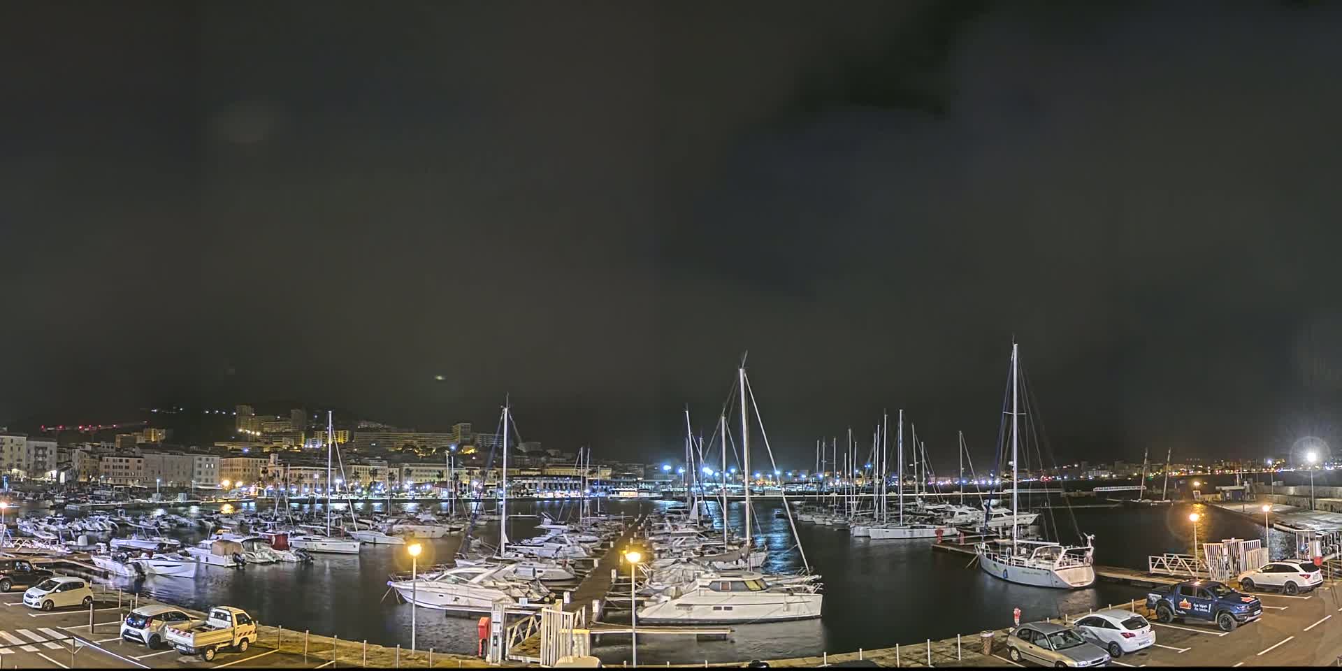 A marina filled with numerous boats and yachts is illuminated at night under a dark, cloudy sky.