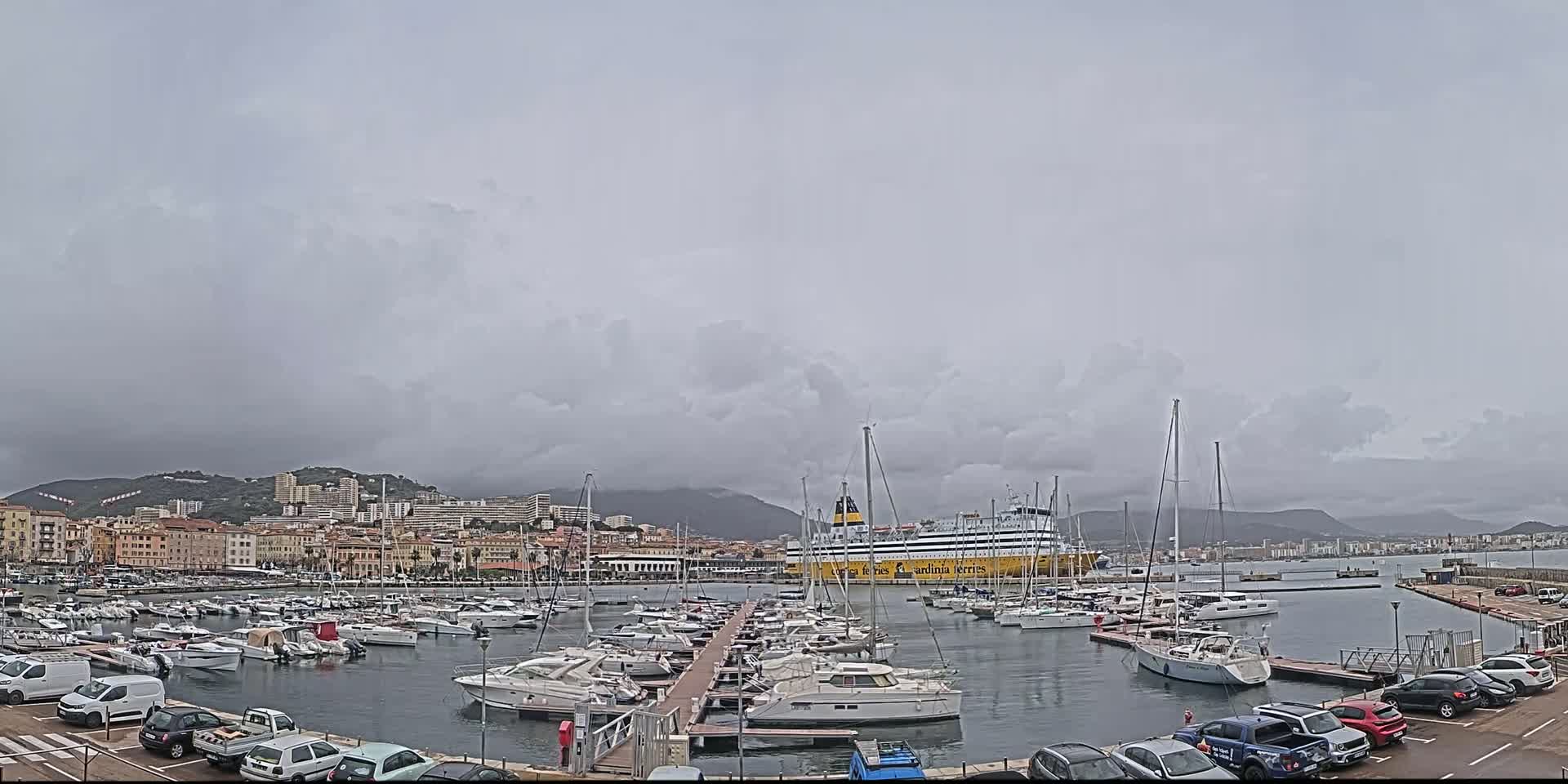 A large ferry is docked in a harbor filled with numerous yachts and sailboats under an overcast sky.