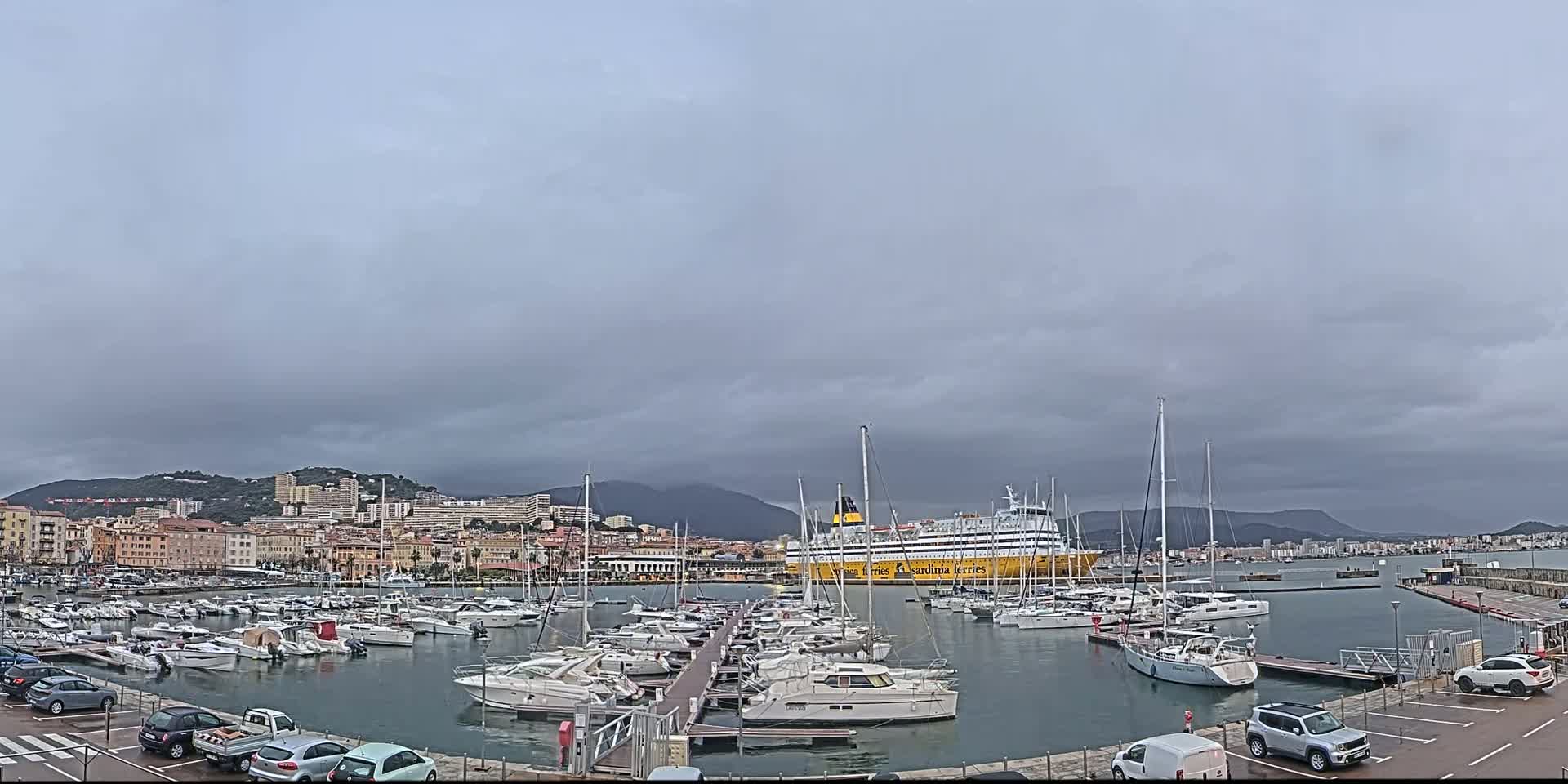 A large ferry is docked in a harbor filled with numerous yachts and sailboats under an overcast sky.