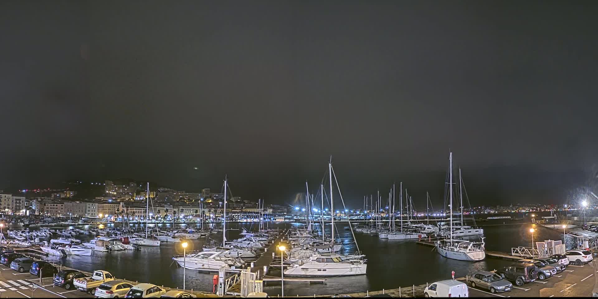 A nighttime view of a marina filled with numerous sailboats and yachts, under a dark, overcast sky.