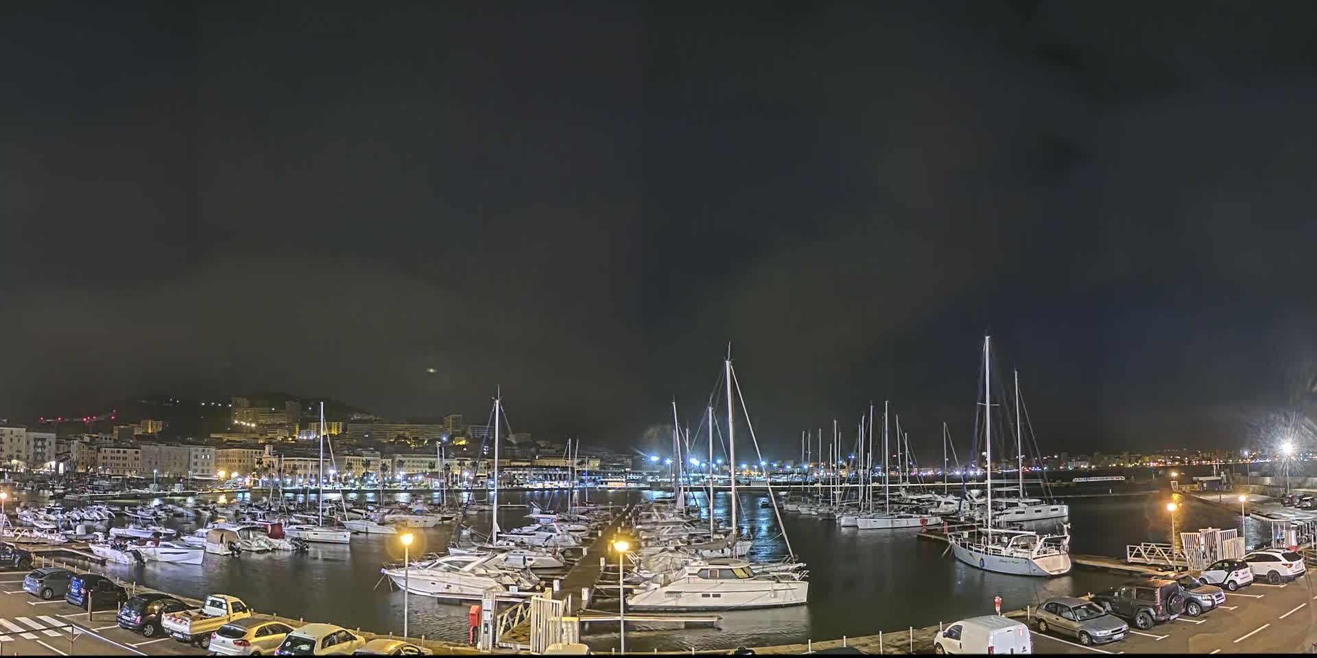 A nighttime harbor filled with numerous boats and yachts, under a dark, cloudy sky, is adjacent to a city illuminated with lights.