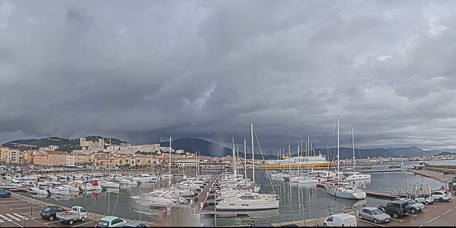 A marina filled with numerous sailboats and motorboats is situated near a city under a heavily overcast sky.