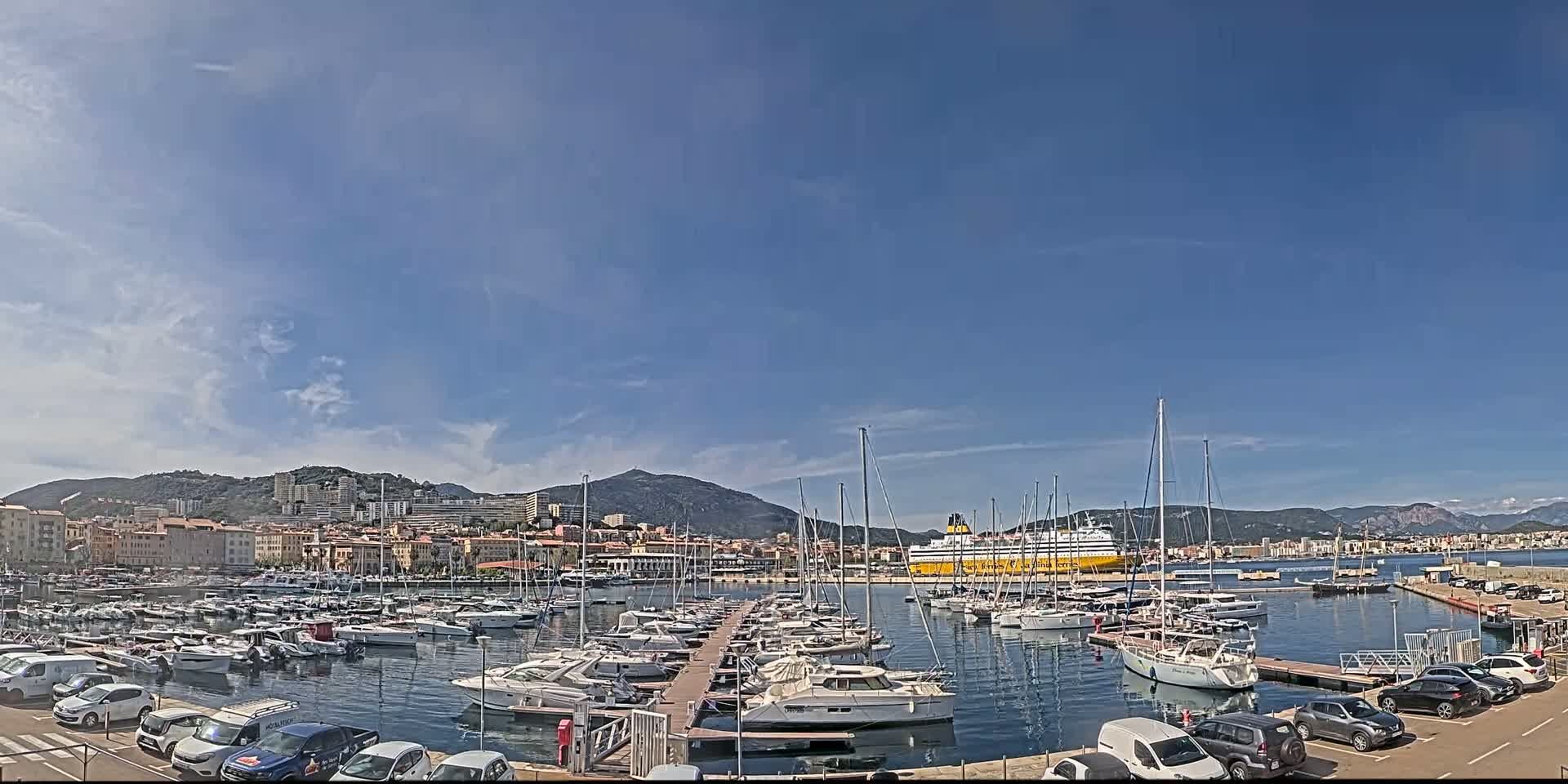 A harbor filled with sailboats and motorboats is situated near a city and mountains under a mostly sunny sky with a few scattered clouds.