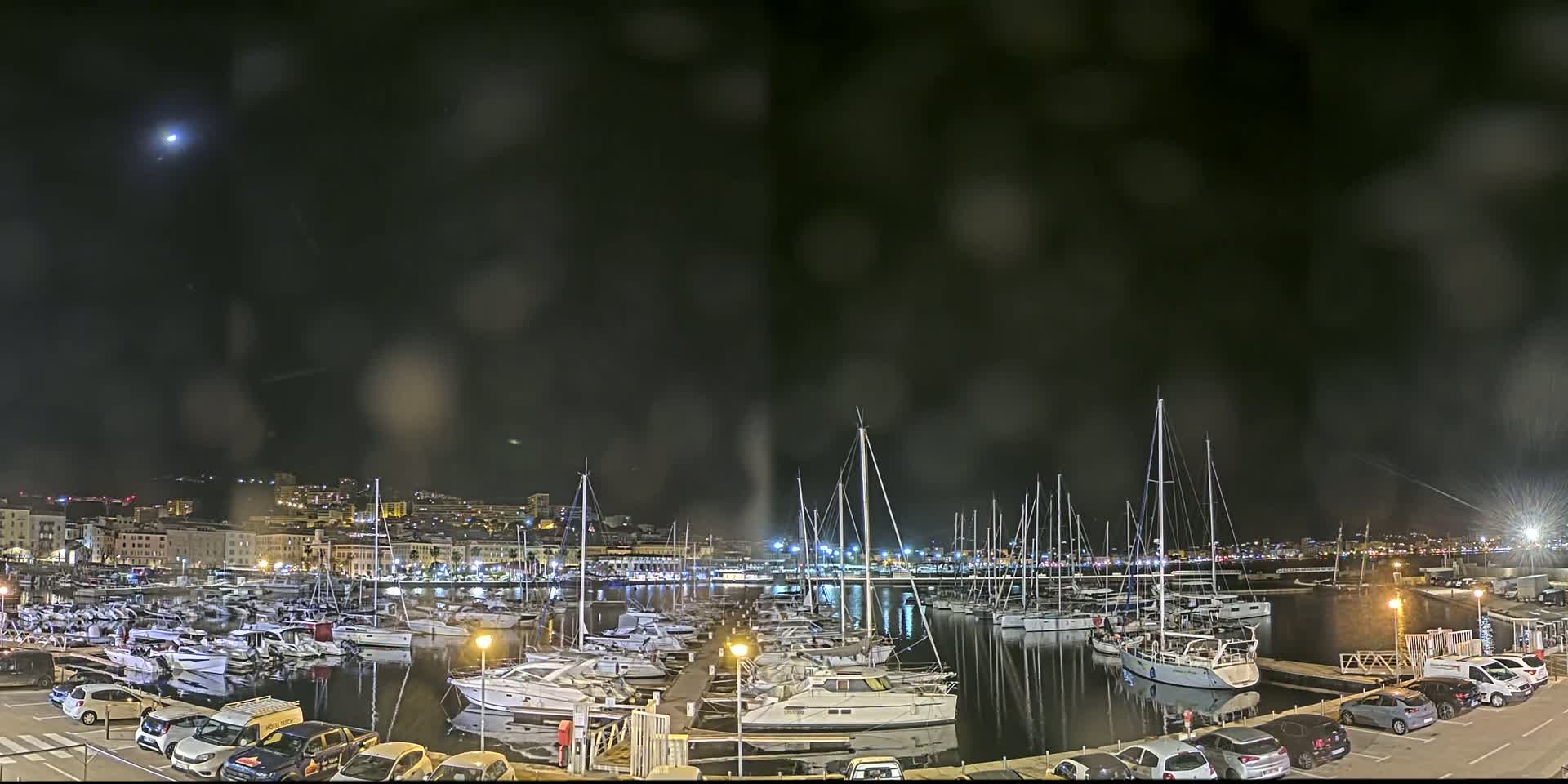 A nighttime view of a harbor filled with boats under a slightly hazy sky, with a moon visible and city lights in the background.