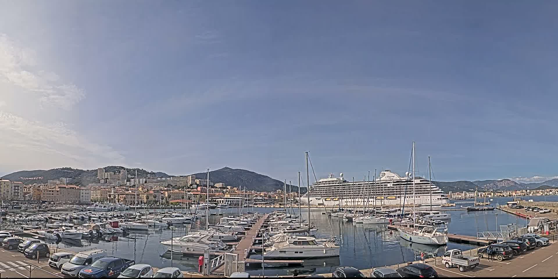 A large cruise ship is docked in a harbor filled with numerous yachts and boats, under a mostly clear sky.