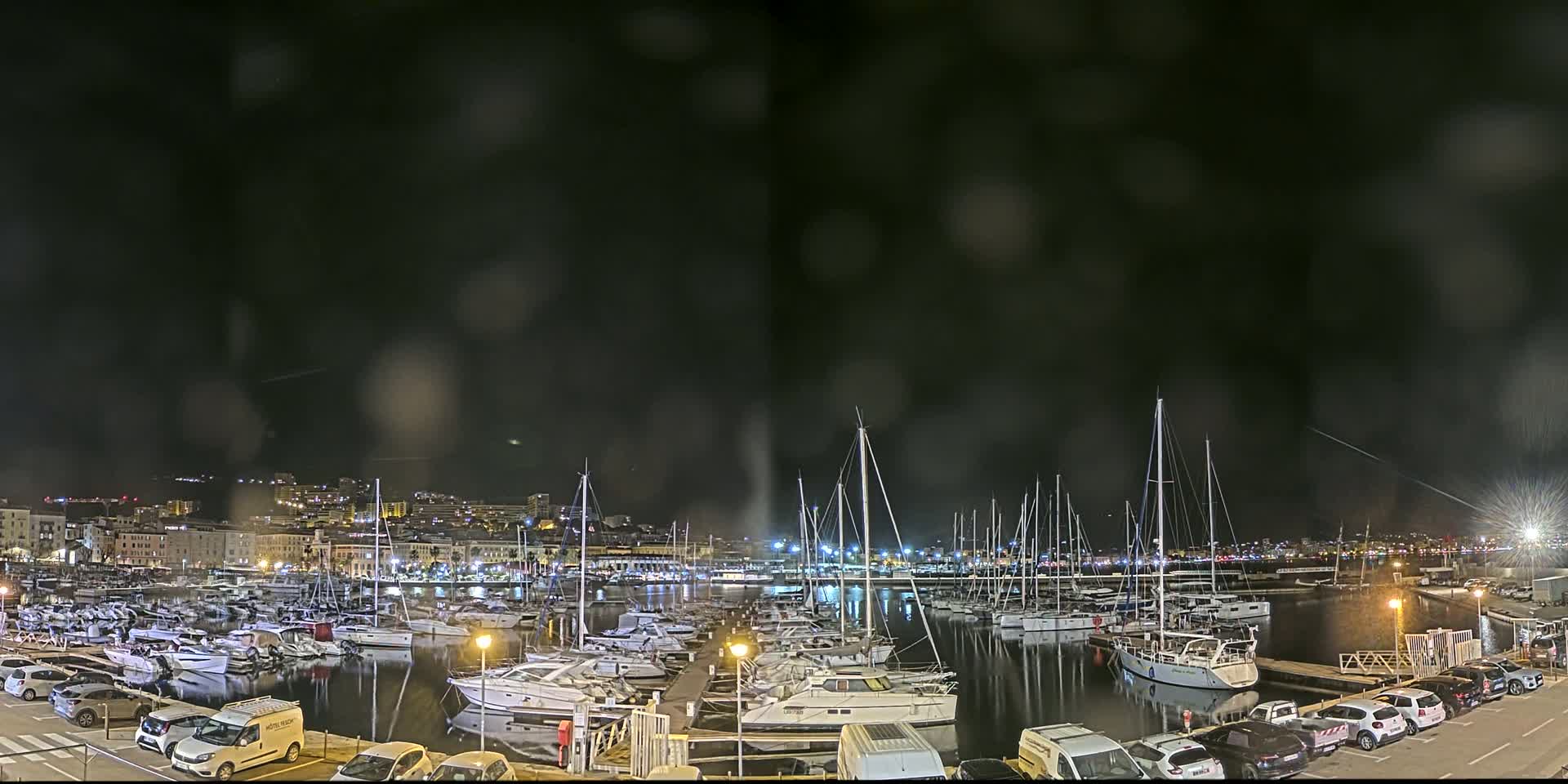 A nighttime view of a harbor filled with sailboats and yachts under a dark, rain-streaked sky.
