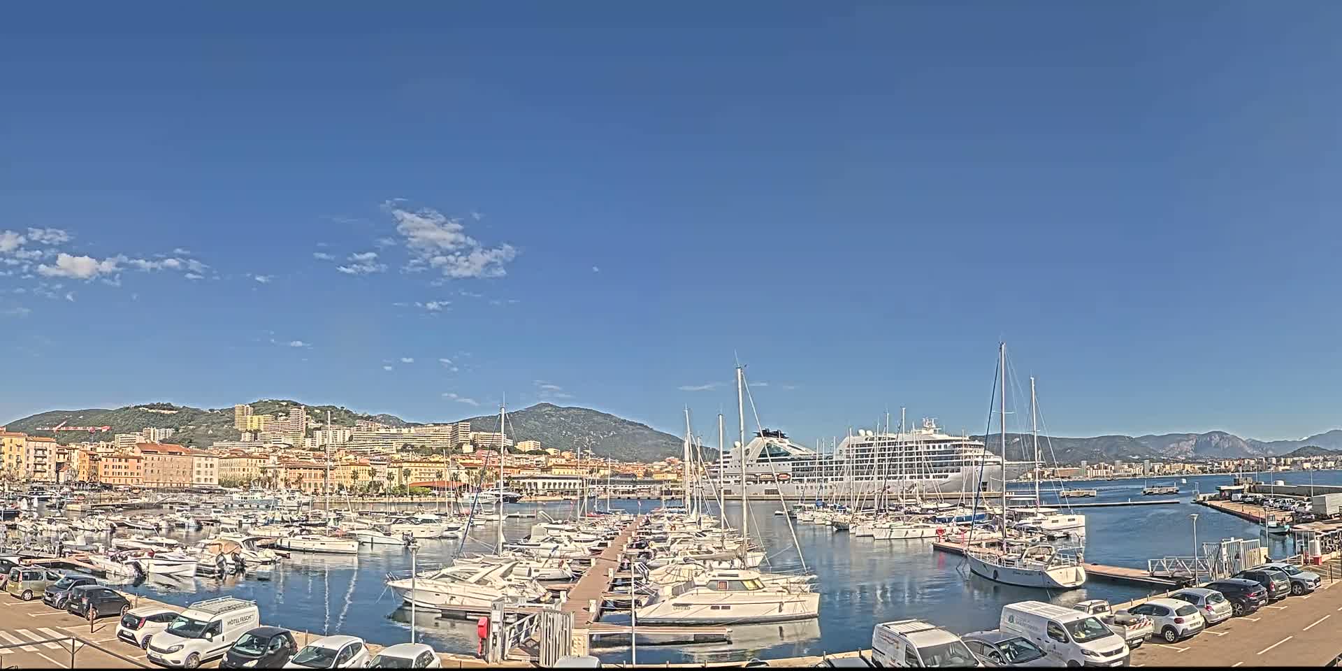 A large cruise ship and many smaller boats are docked in a harbor, with a city and mountains visible in the background under a mostly clear blue sky.