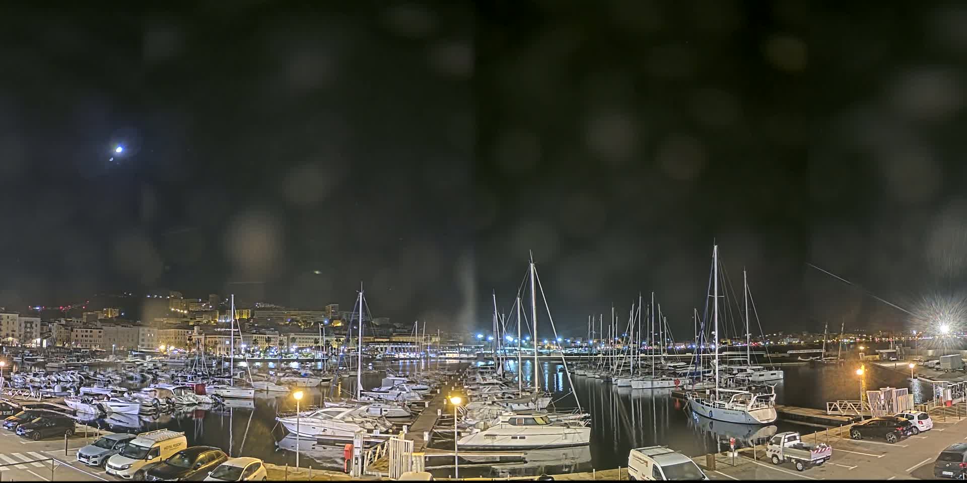 A nighttime view of a marina filled with docked sailboats and motorboats under a dark, slightly hazy sky, with a faint moon visible.