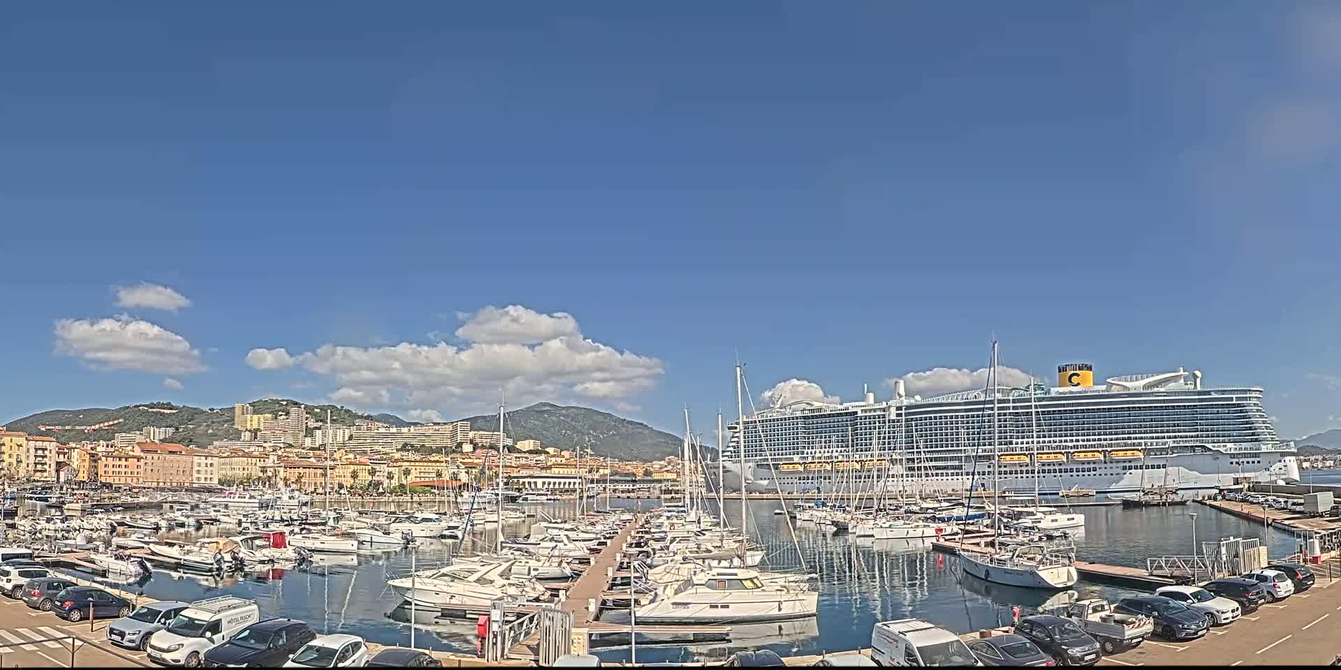 A large cruise ship is docked in a harbor filled with smaller boats, under a mostly sunny sky with some scattered clouds, near a city nestled against a mountain range.