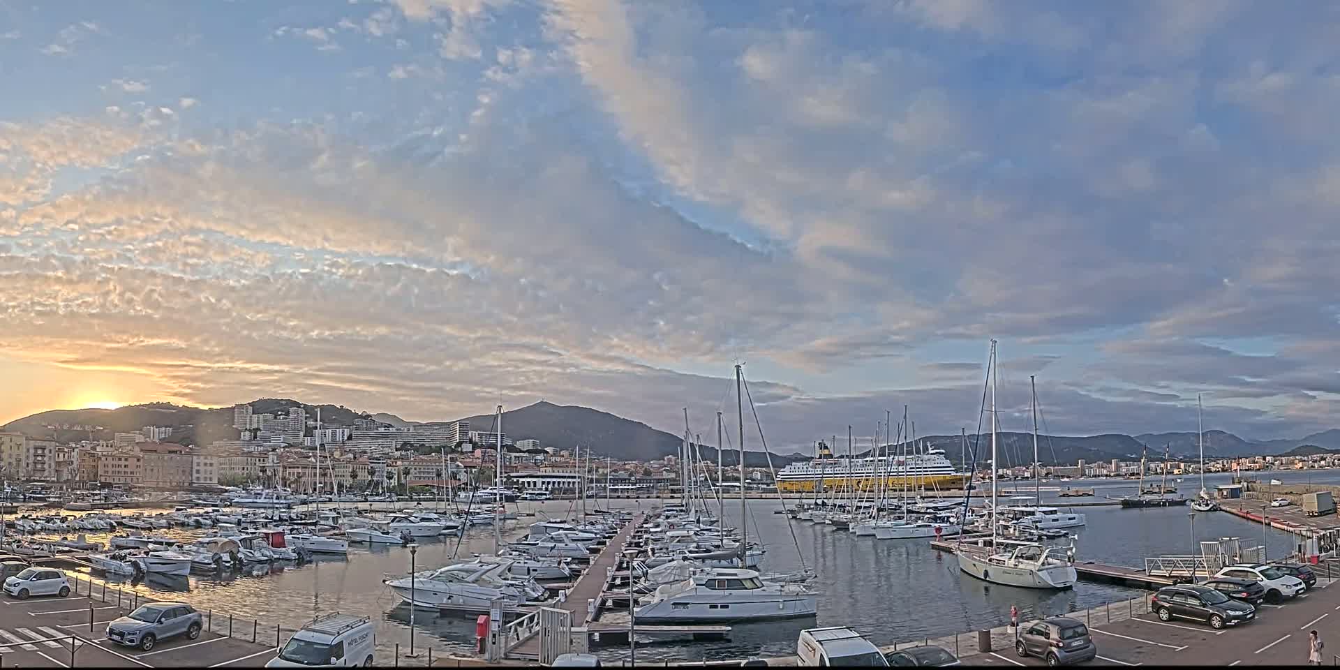 A harbor filled with sailboats and yachts is nestled beside a city at sunset under a partly cloudy sky.