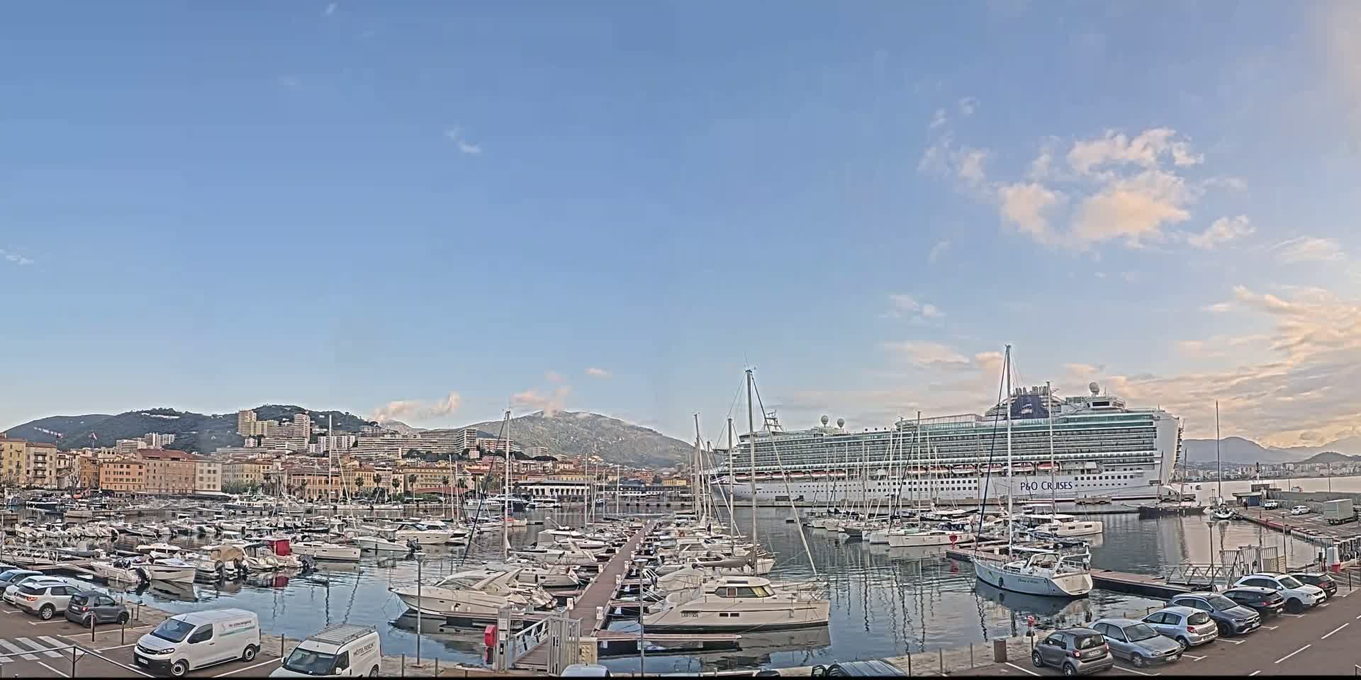A large cruise ship is docked in a harbor filled with numerous smaller boats, under a mostly clear sky with a few clouds.