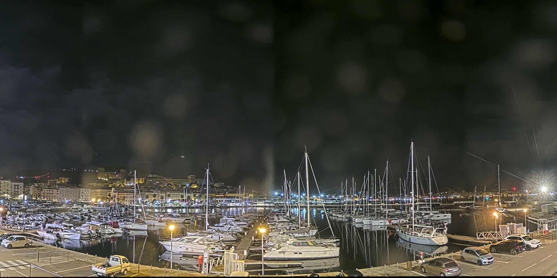 A nighttime view of a marina filled with numerous boats under a dark, cloudy sky with light rain.