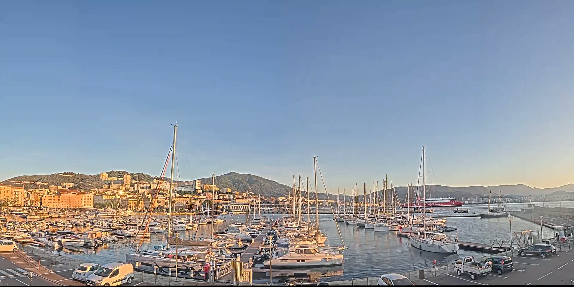 Under a clear blue sky, a harbor filled with sailboats and motorboats is nestled next to a city backed by mountains.