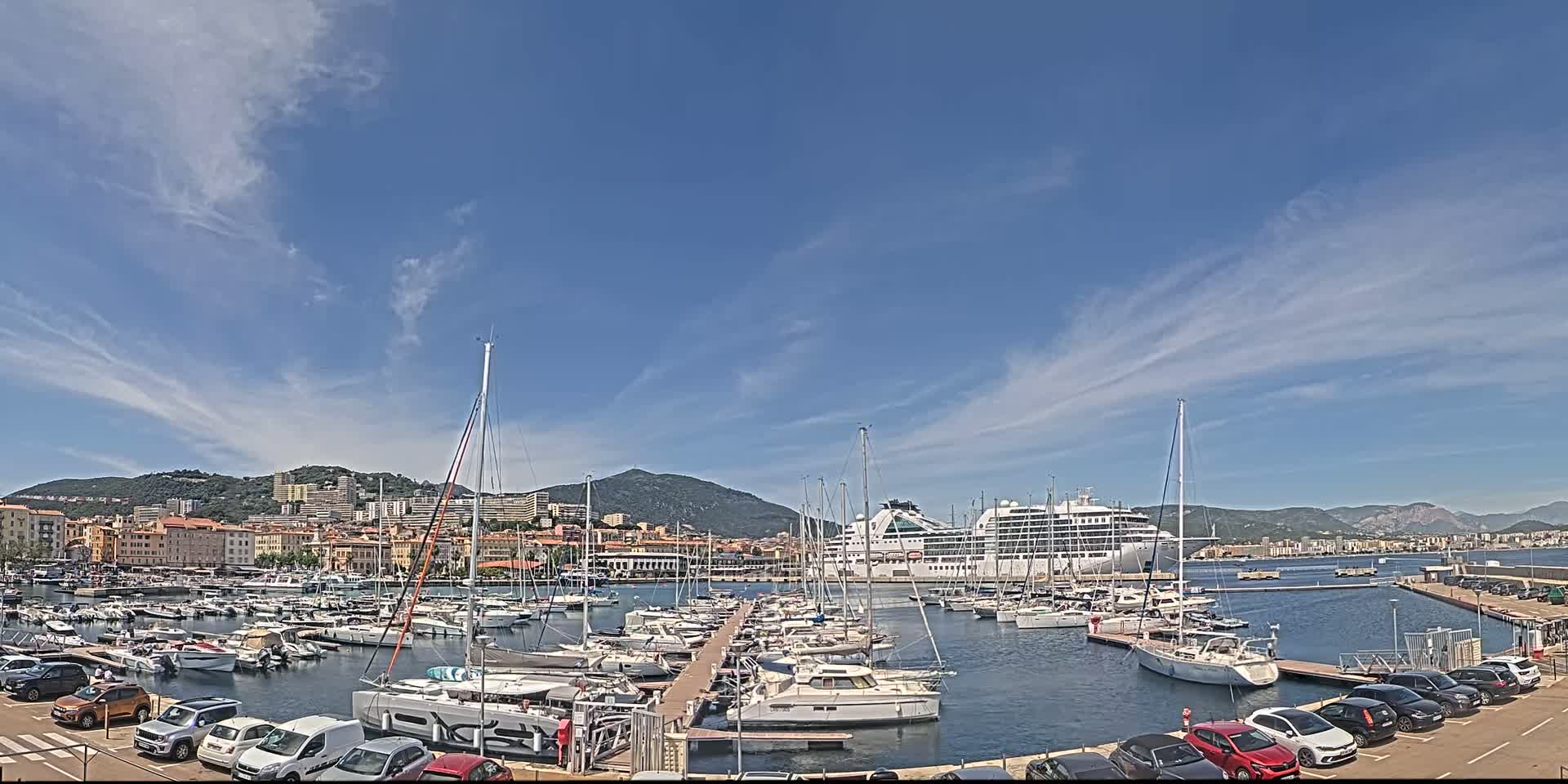 A harbor filled with numerous yachts and a large cruise ship is nestled against a backdrop of a town and mountains under a mostly sunny sky with scattered clouds.