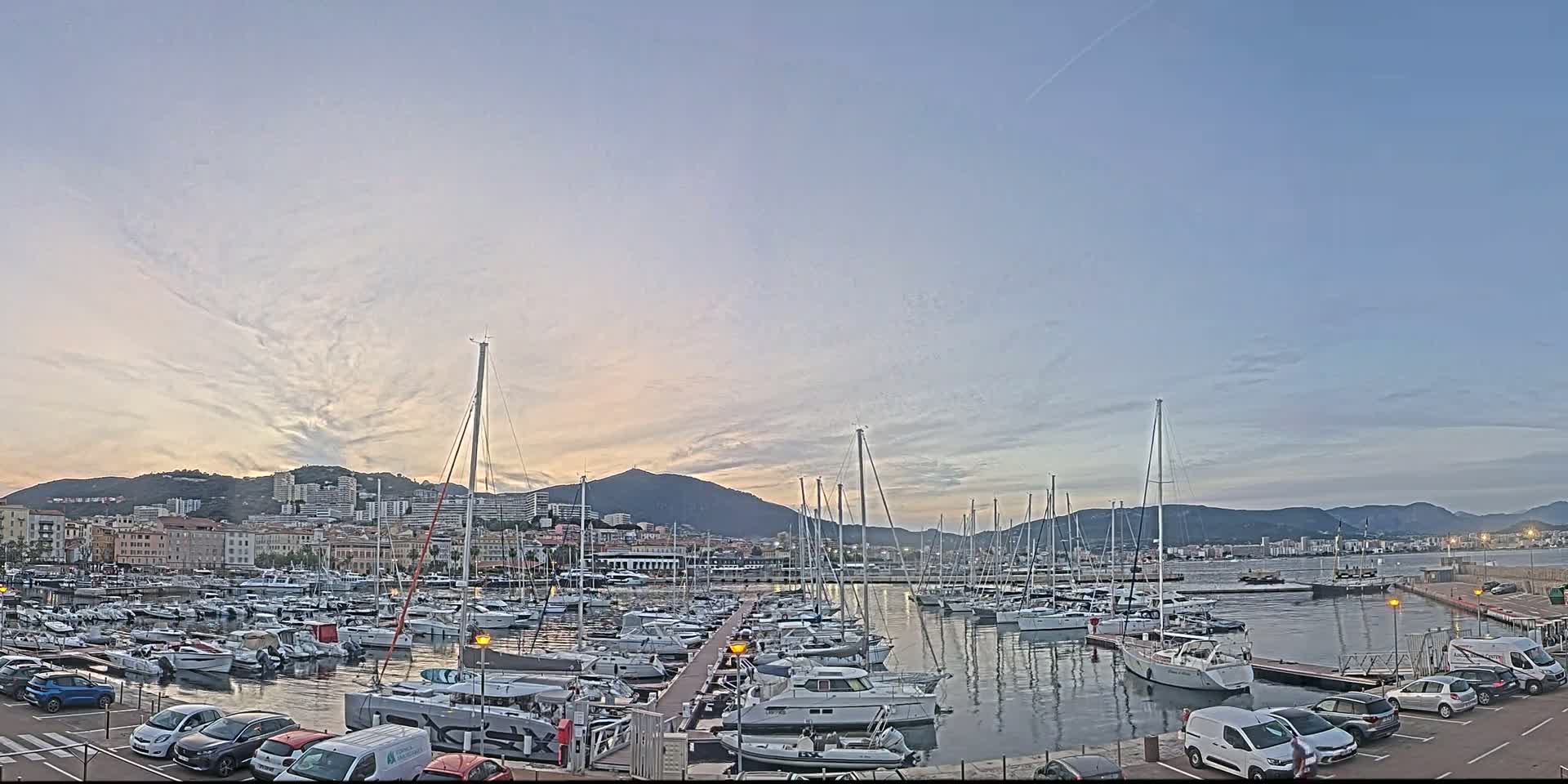 A marina filled with numerous boats is situated in front of a city at sunset under a mostly clear sky.