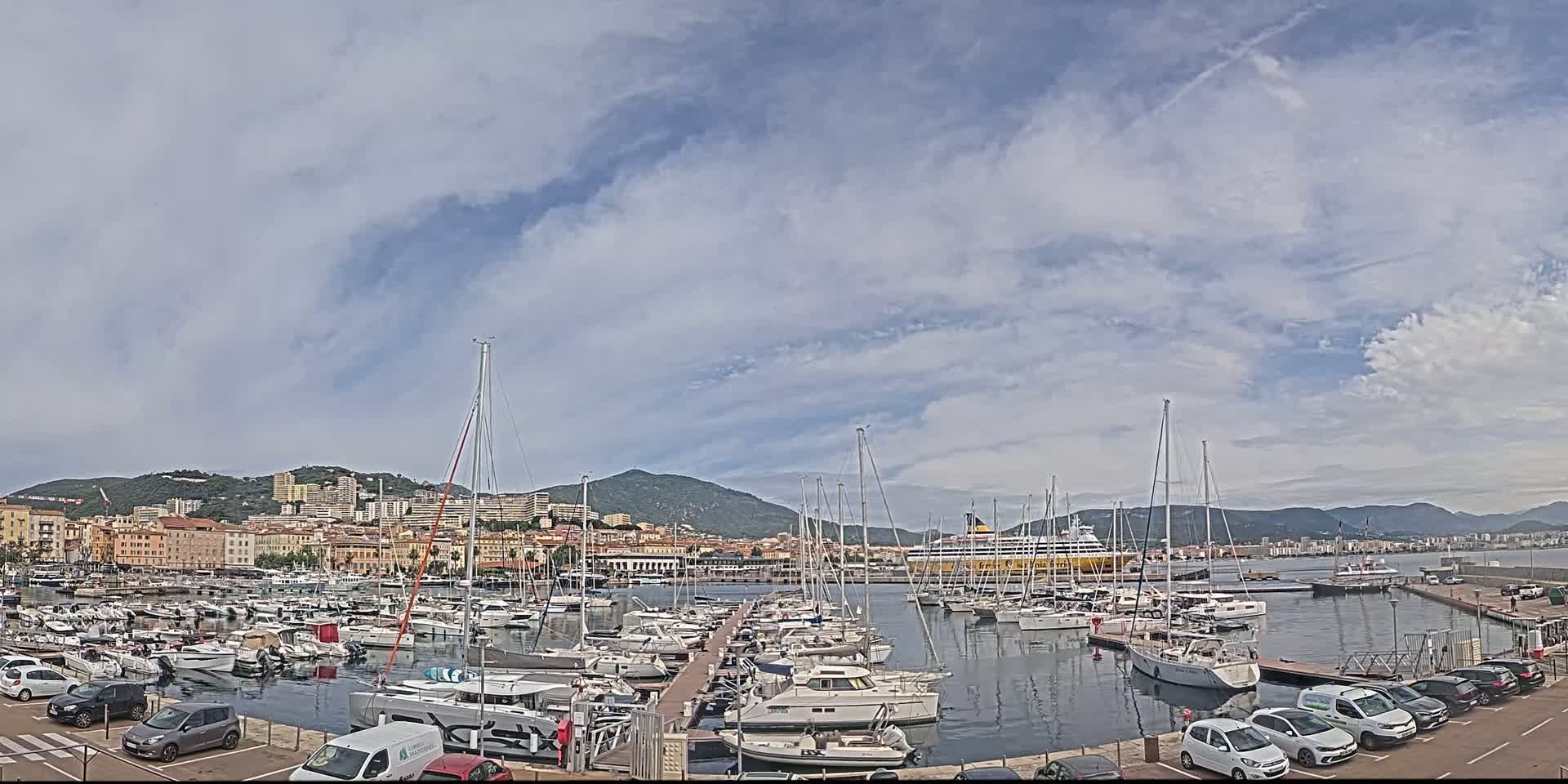 A marina filled with sailboats and yachts is situated in front of a city nestled against a mountain range under a partly cloudy sky.
