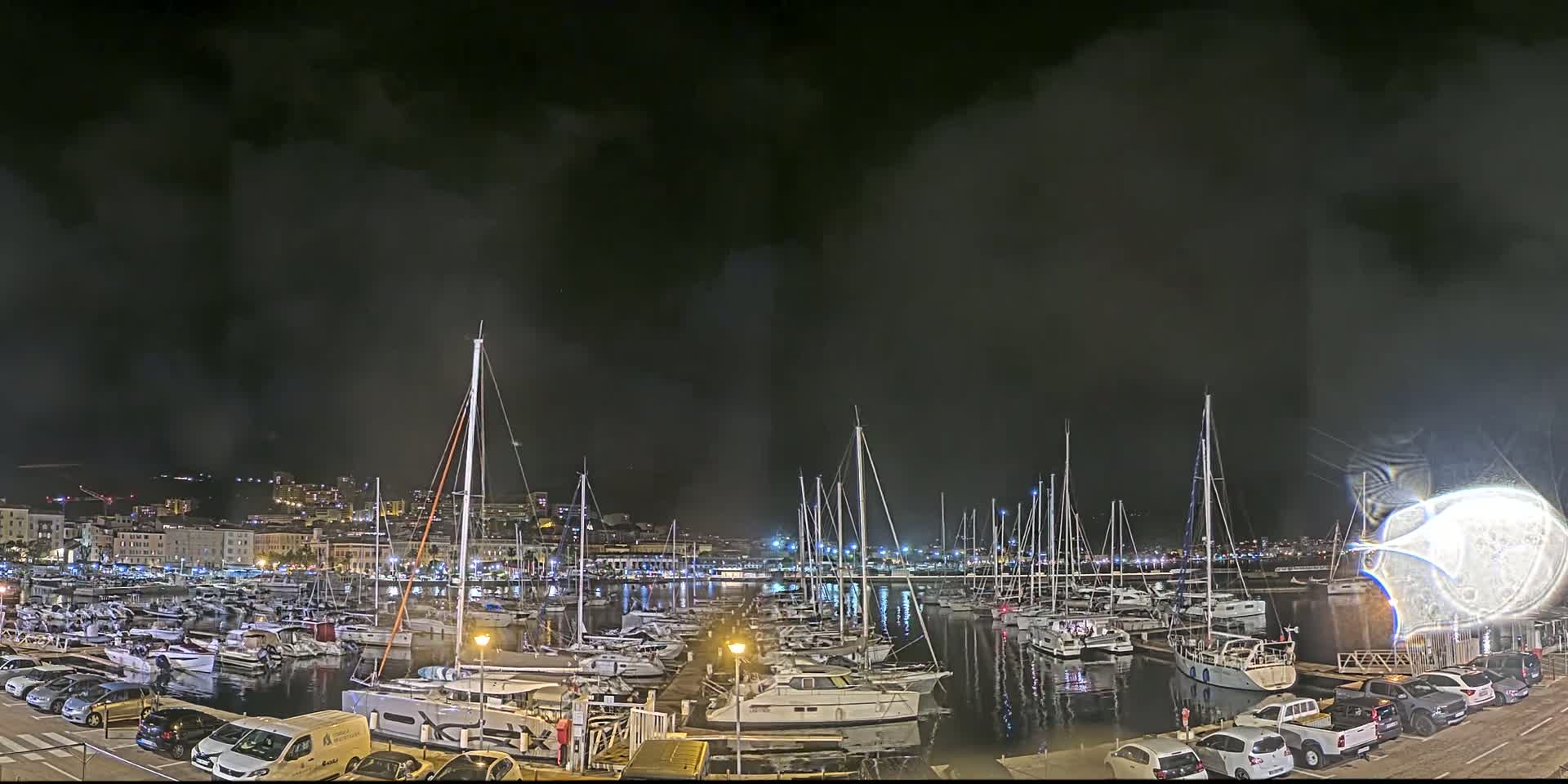 A nighttime harbor filled with many boats, under a cloudy sky, with a city skyline in the background.