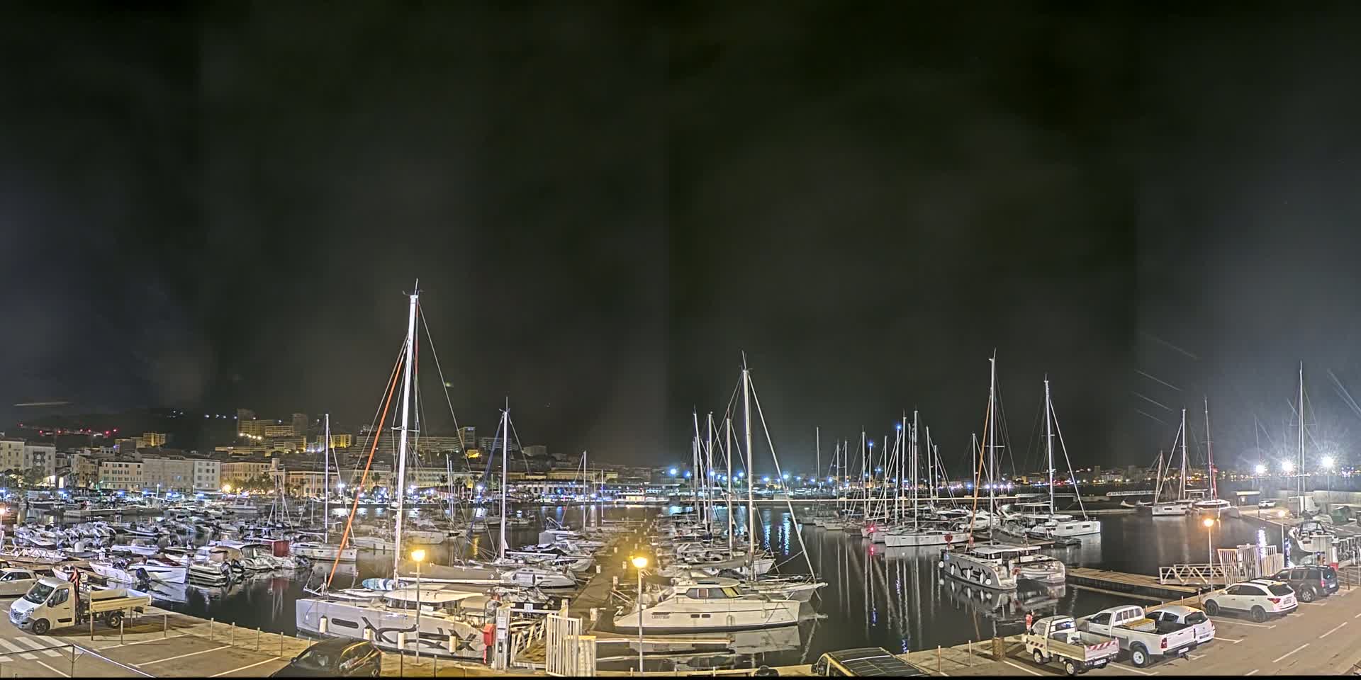 A marina filled with sailboats and motorboats is illuminated at night under a dark, cloudy sky.