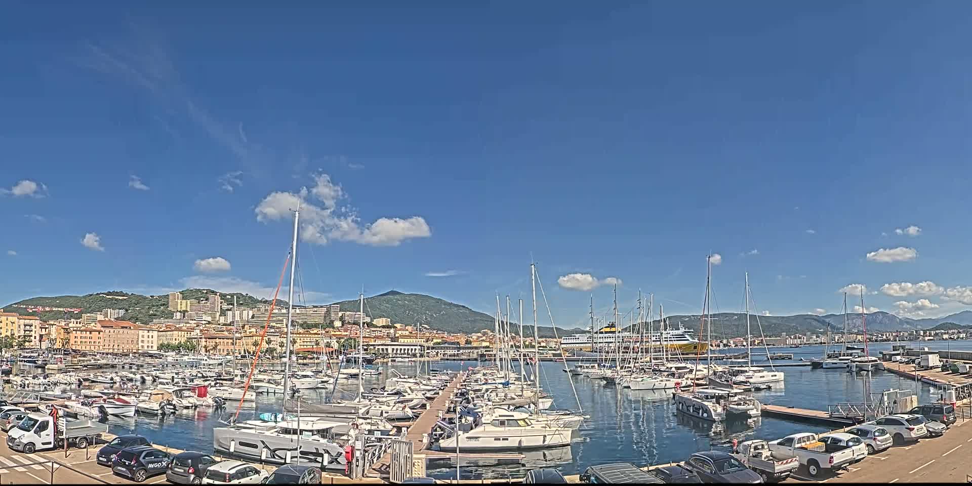 A marina filled with sailboats and motorboats is situated in front of a town and mountains under a mostly sunny sky with some scattered clouds.