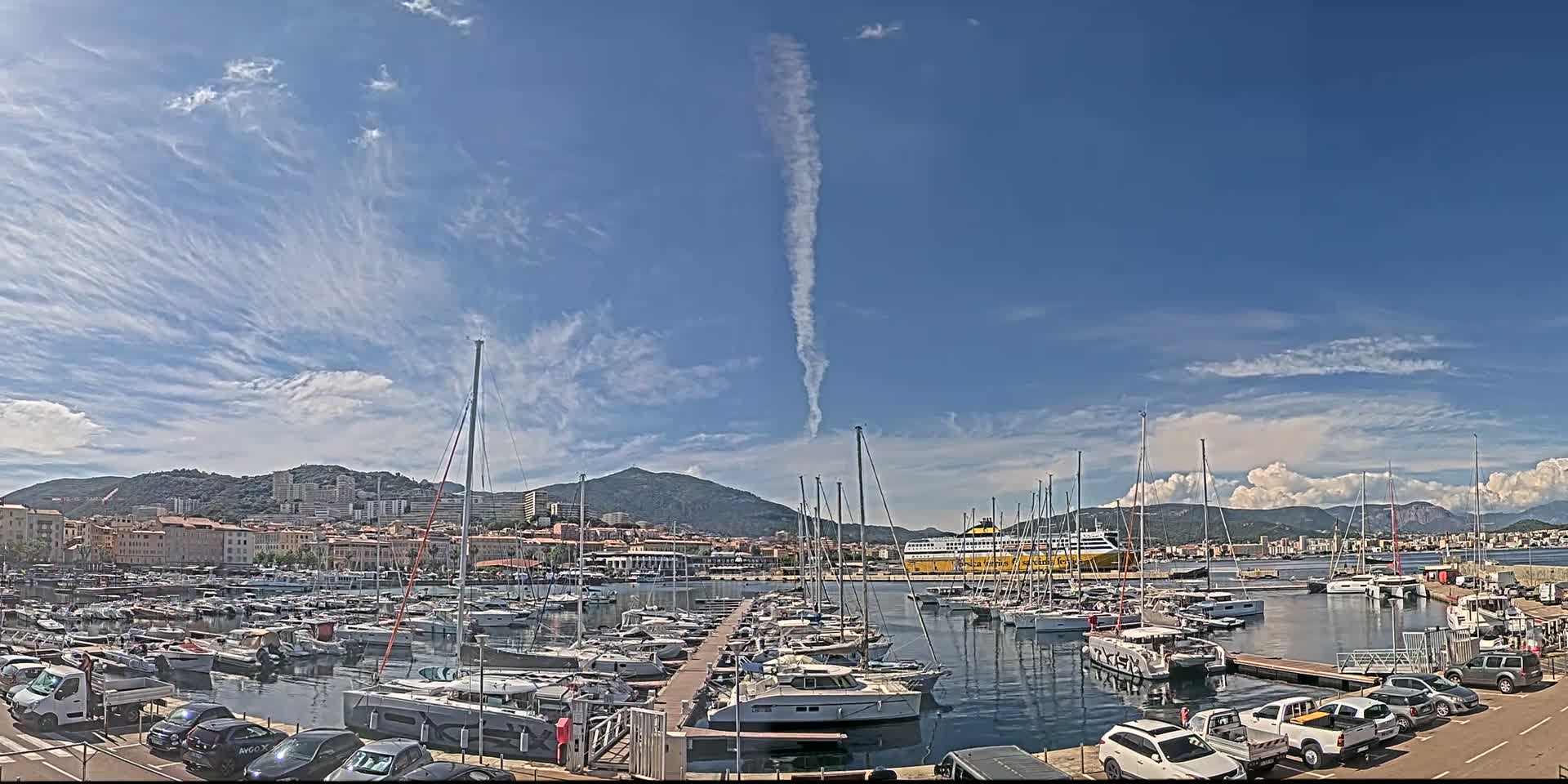 A marina filled with sailboats and yachts is situated in front of a city nestled against mountains under a mostly sunny sky with scattered clouds and a single contrail.