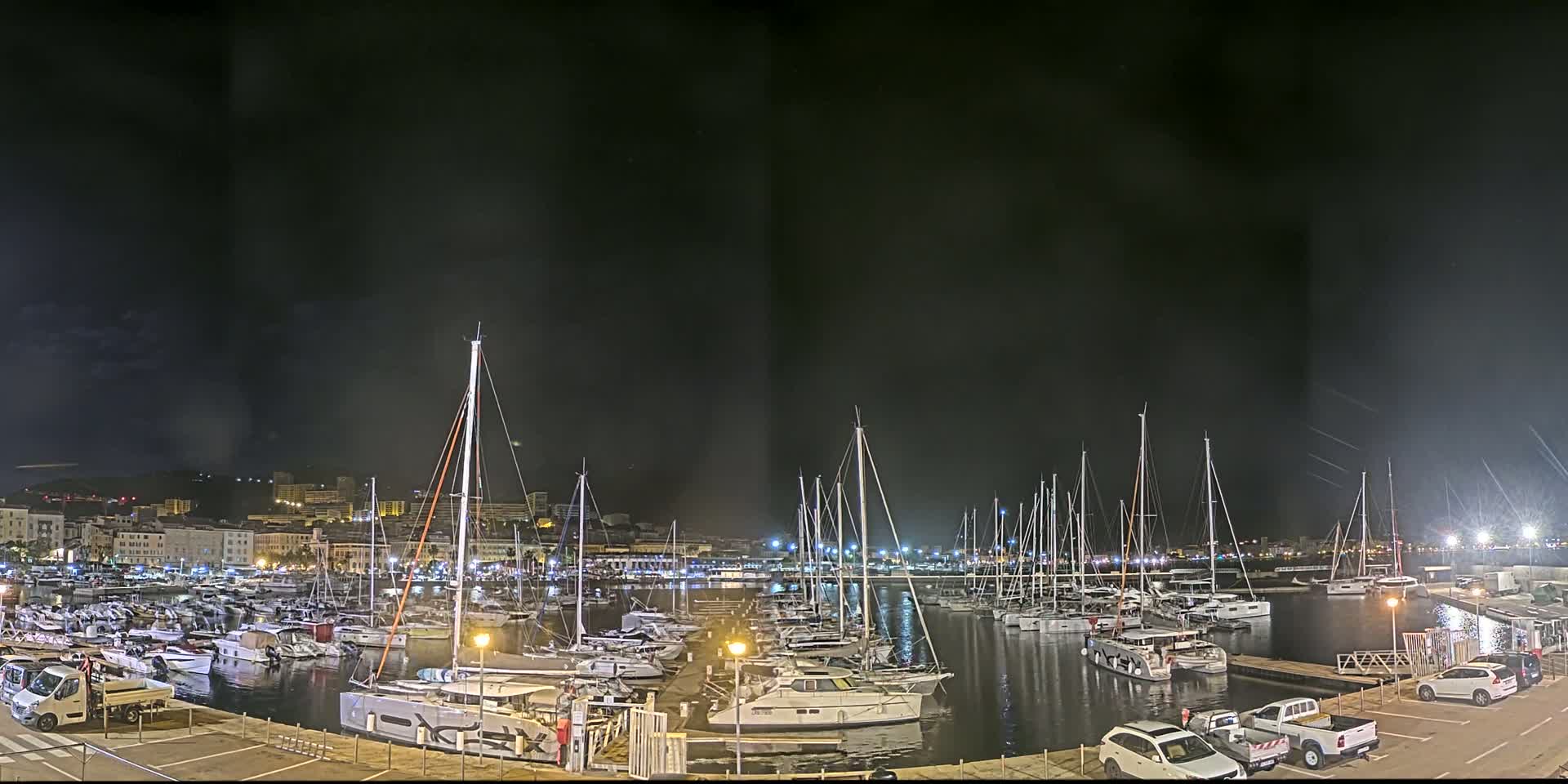 A marina filled with numerous sailboats and motorboats is illuminated at night under a dark, possibly overcast sky.