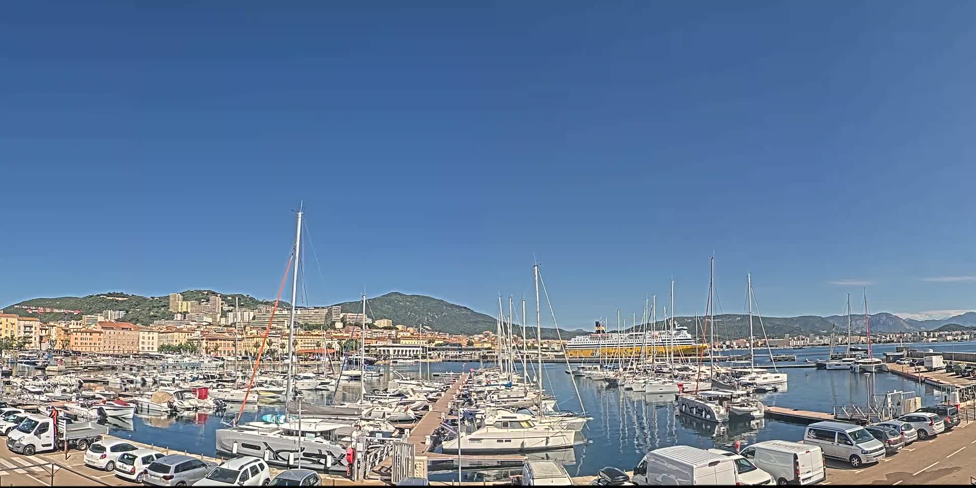 A marina filled with sailboats and motorboats is situated in front of a town nestled against a mountain range under a clear blue sky.