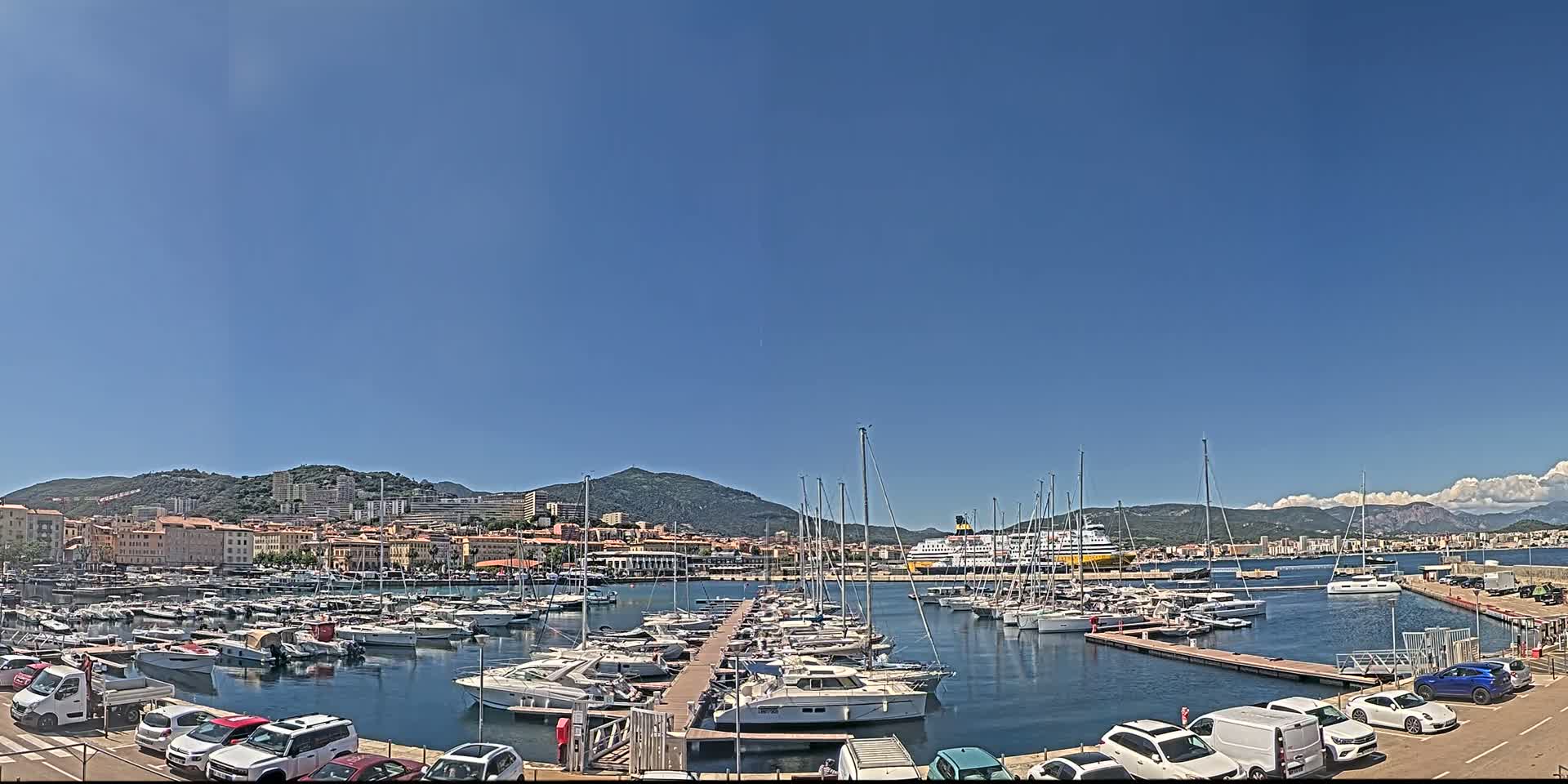 A marina filled with numerous boats and yachts is situated in front of a town nestled against a mountain range under a clear, sunny sky.