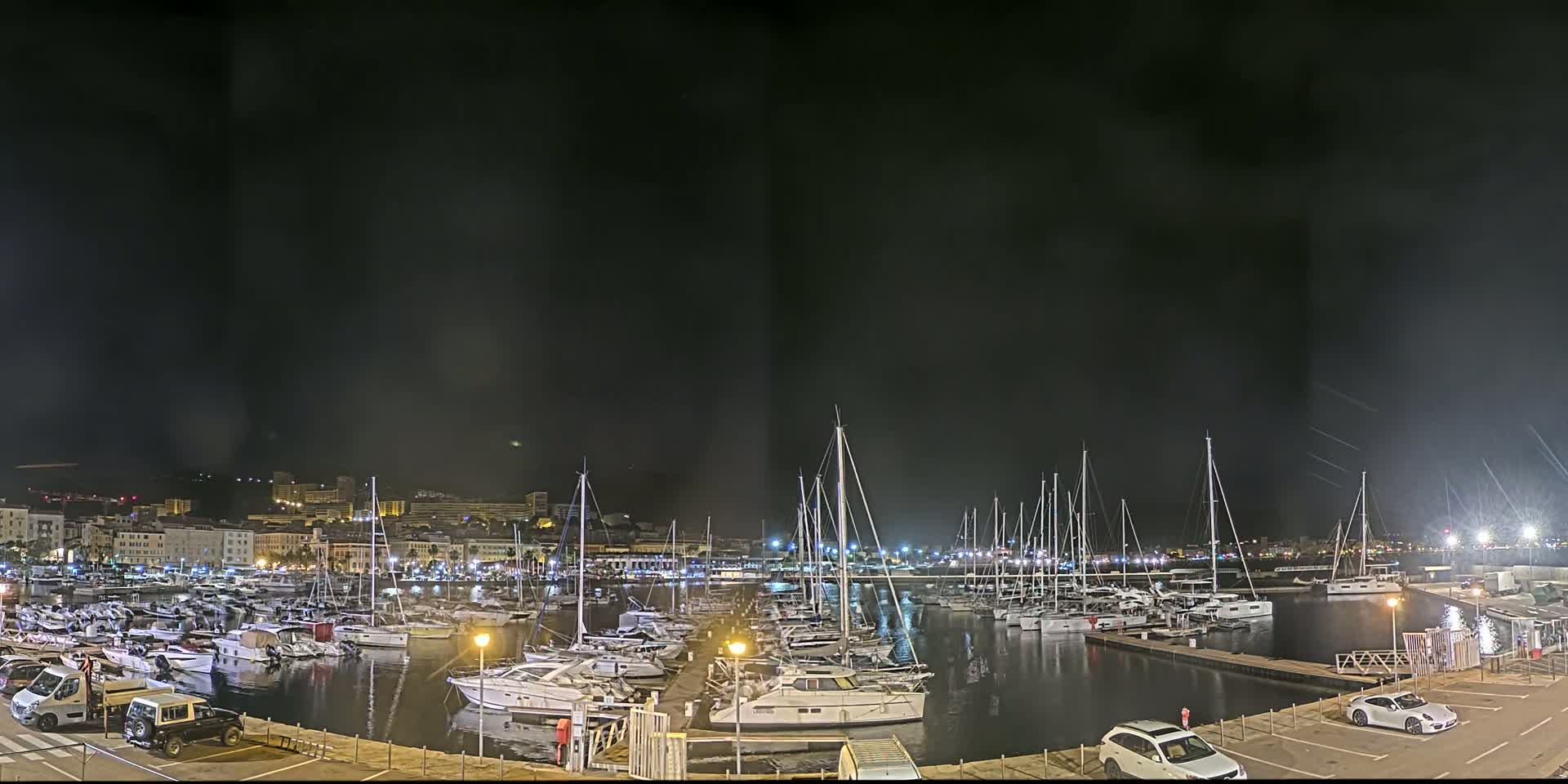 A nighttime view of a marina filled with sailboats and yachts, under a dark, possibly slightly hazy sky.