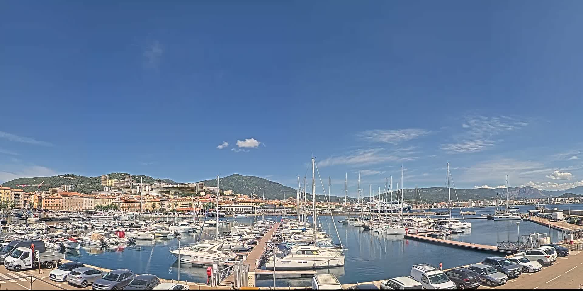 A harbor filled with numerous sailboats and motorboats is situated in front of a city nestled against mountains under a mostly clear, sunny sky.