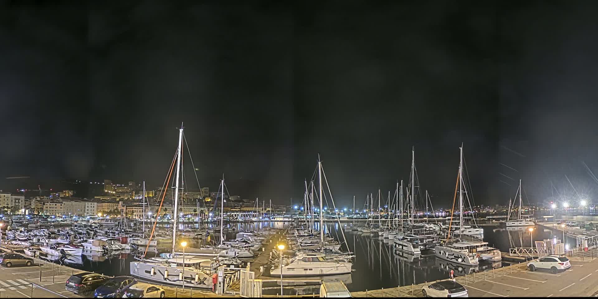 A nighttime view of a marina filled with numerous sailboats and yachts, under a dark, possibly cloudy sky.