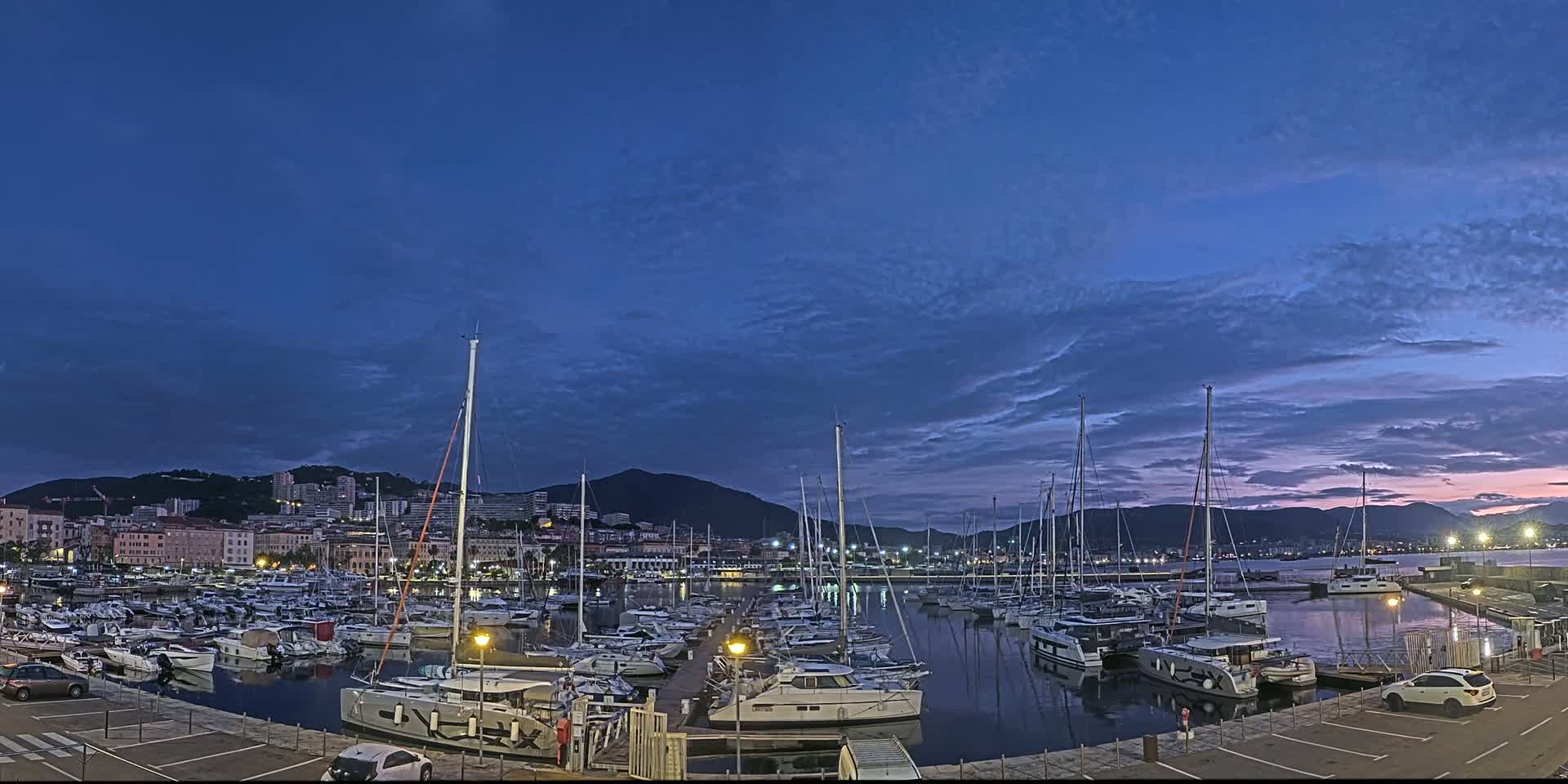 A marina filled with sailboats and motorboats is situated at twilight under a partly cloudy sky, with a city and mountains in the background.