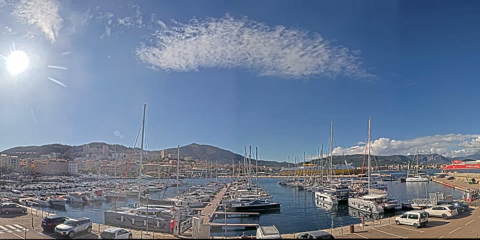 A sunny day reveals a harbor filled with numerous sailboats and motorboats docked near a city backed by mountains.
