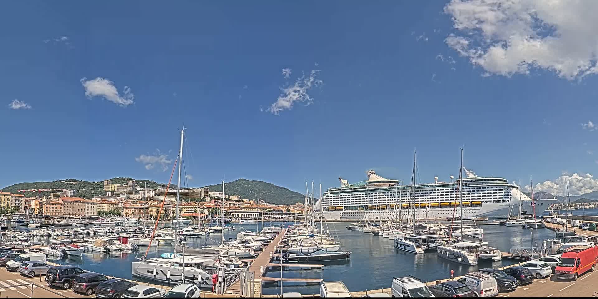 A large cruise ship is docked in a harbor filled with numerous yachts and sailboats, under a mostly sunny sky with a few scattered clouds.