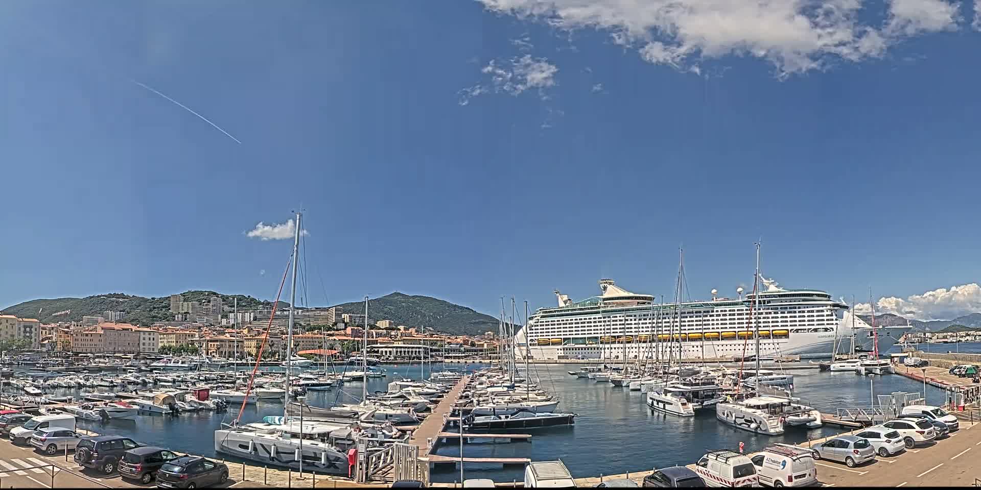 A large cruise ship is docked in a harbor filled with numerous smaller boats under a mostly sunny sky.