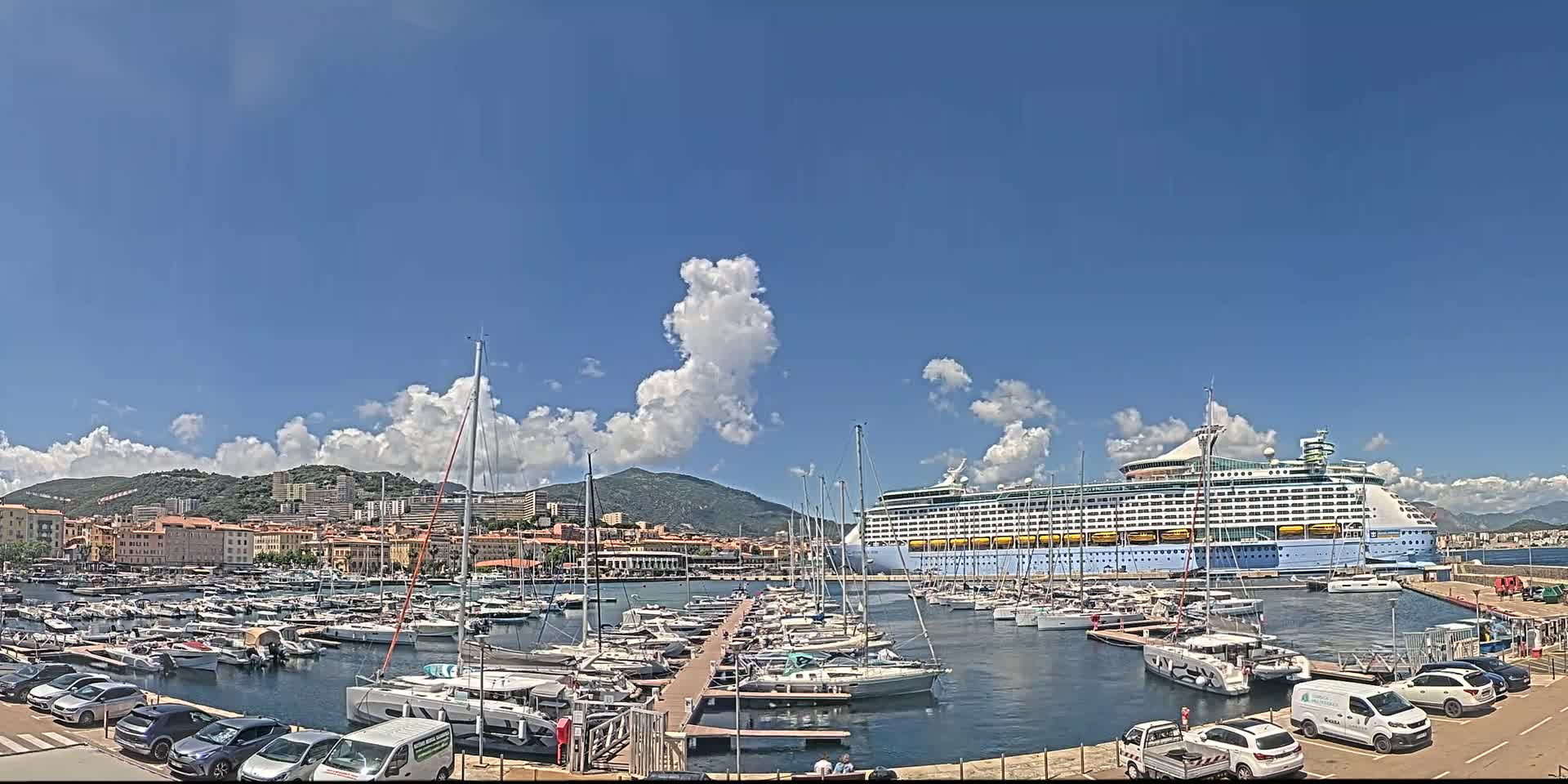 A large cruise ship is docked in a harbor filled with many smaller boats, under a mostly sunny sky with scattered cumulus clouds.