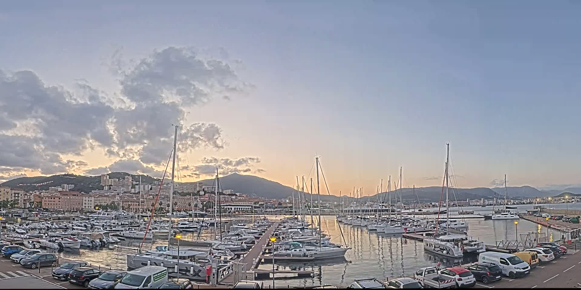 A marina filled with sailboats and yachts is situated near a city at sunset under a partly cloudy sky.