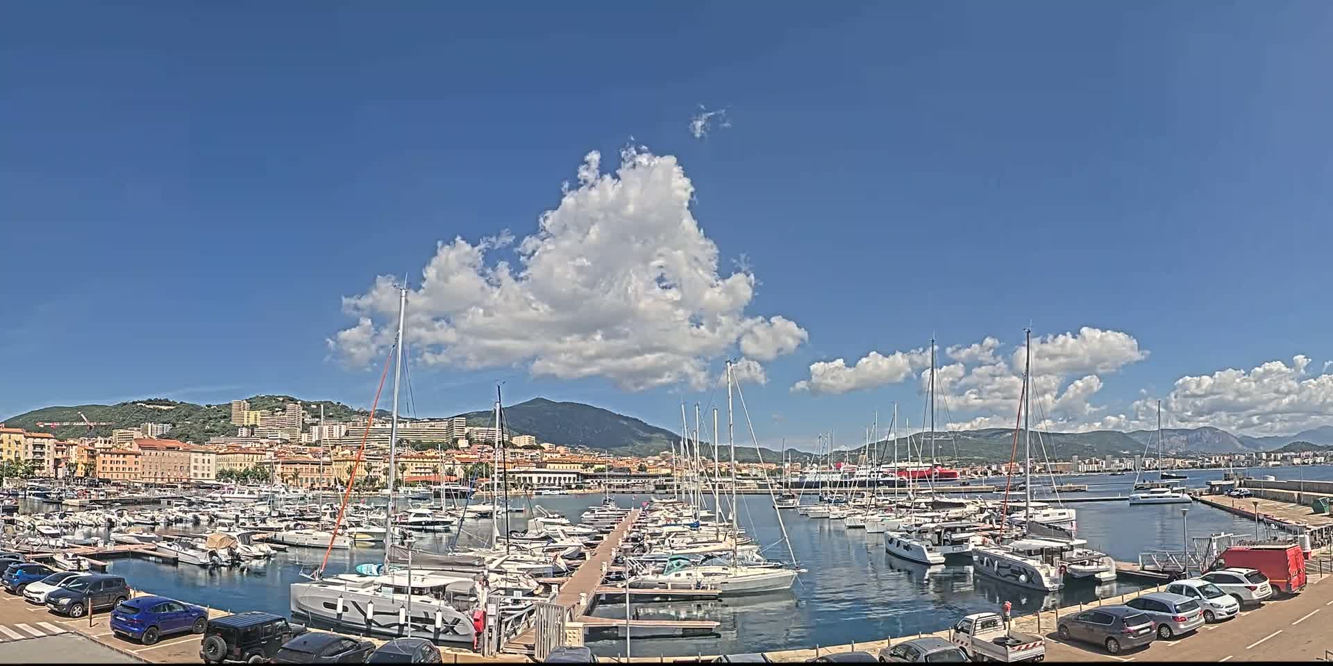 A marina filled with numerous sailboats and motorboats is situated in front of a city nestled against a mountain range under a mostly sunny sky with some puffy cumulus clouds.