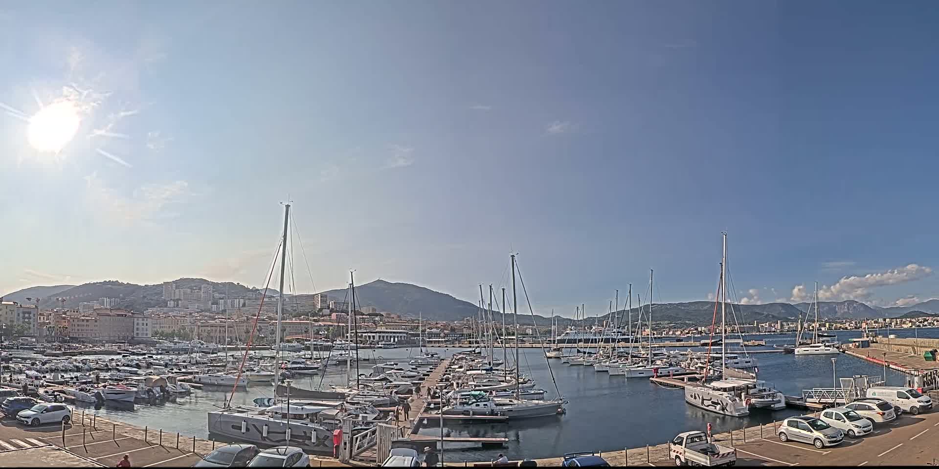 A sunny day shows a marina filled with sailboats and motorboats, with a city and mountains in the background.