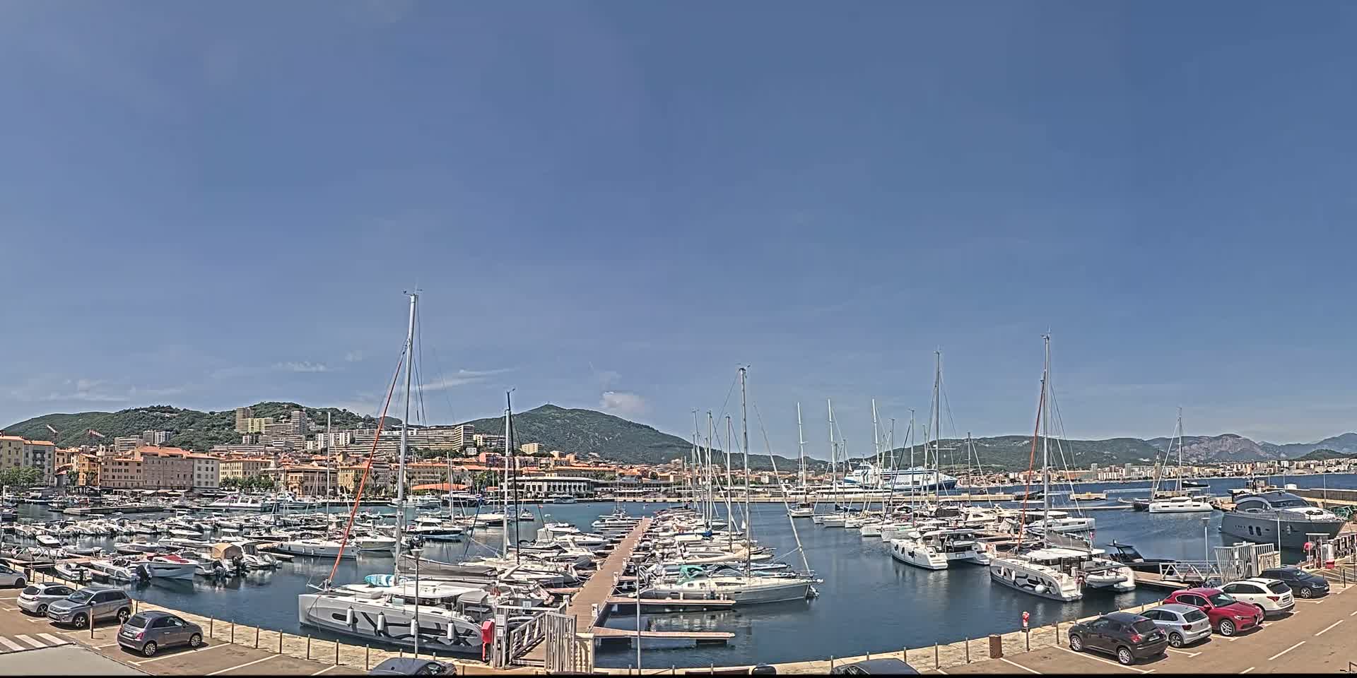 A marina filled with sailboats and yachts is situated near a town nestled against a hillside under a clear blue sky.