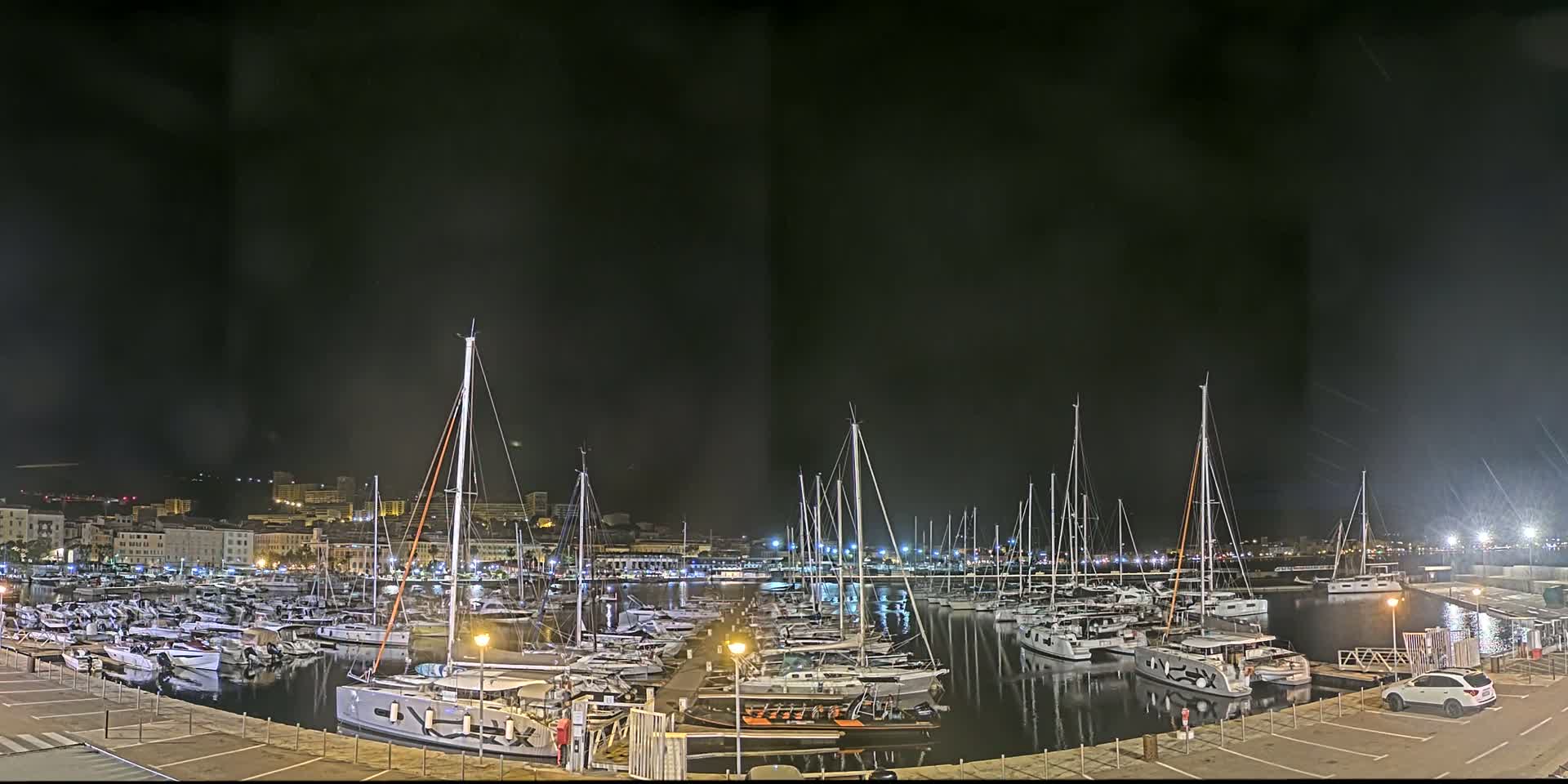 A marina at night, filled with numerous sailboats and motorboats, is illuminated under a dark sky.
