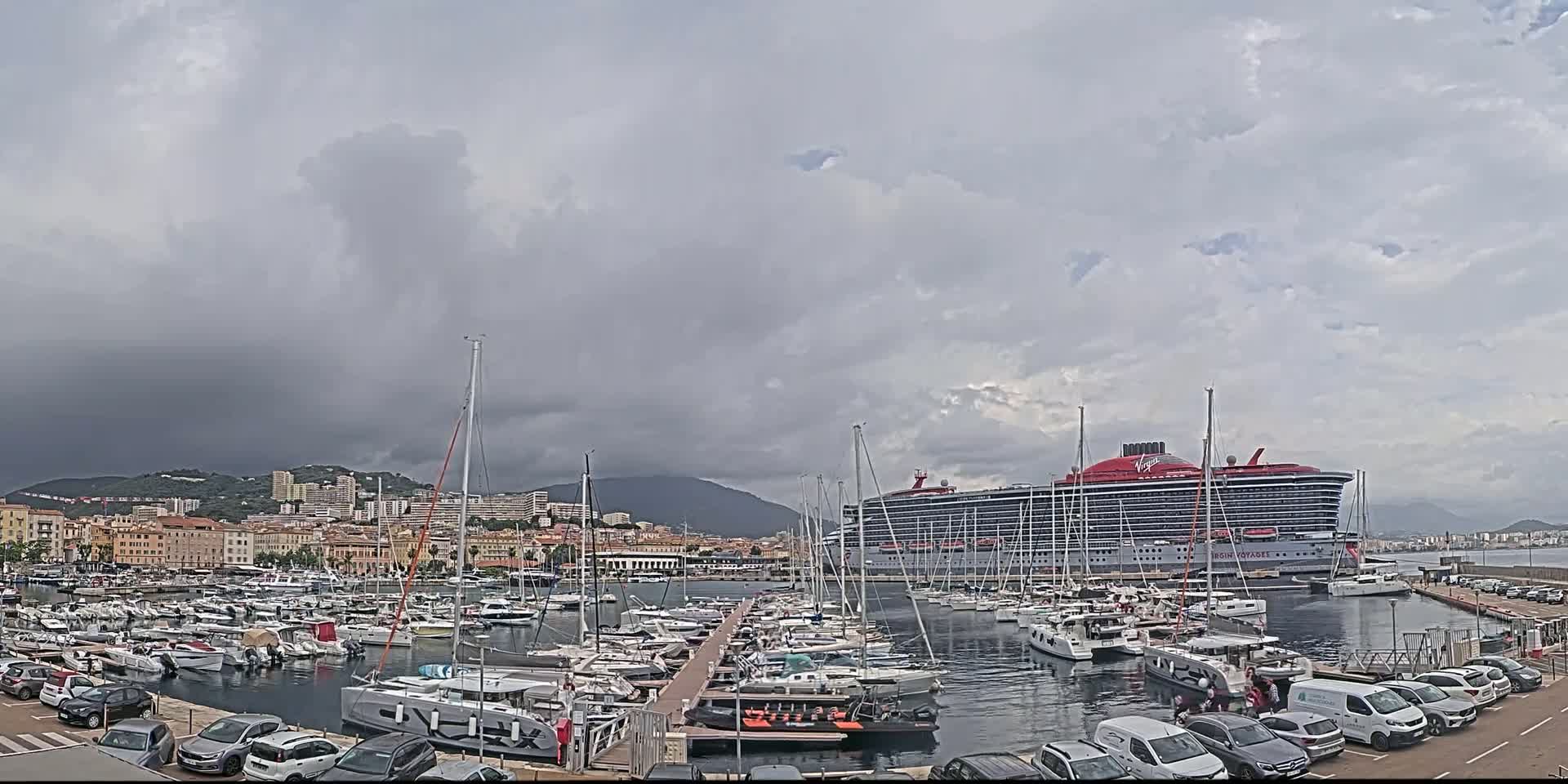 A large cruise ship is docked in a harbor filled with numerous smaller boats under a cloudy sky.
