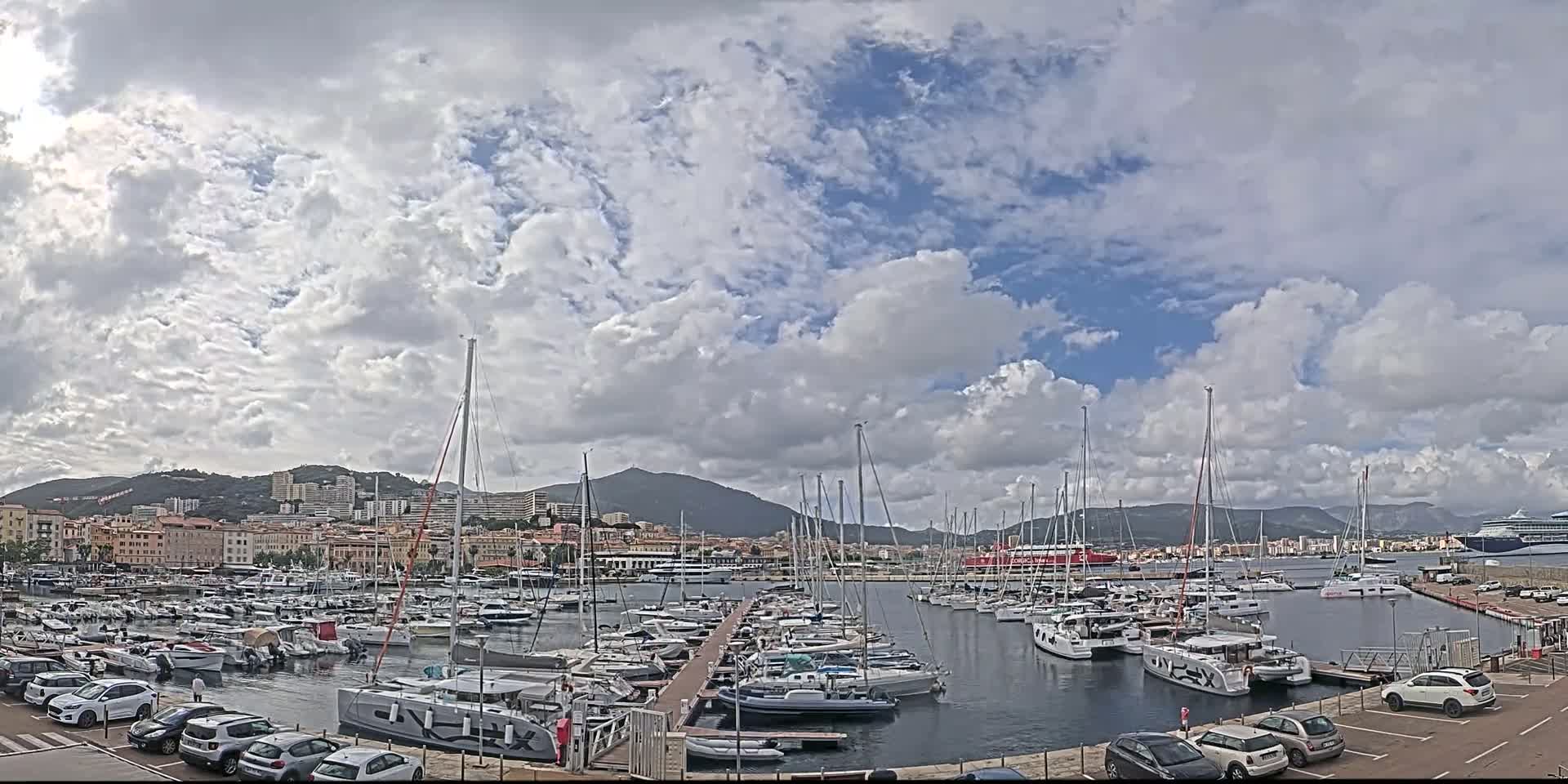 A harbor filled with numerous sailboats and yachts is nestled against a cityscape under a partly cloudy sky.