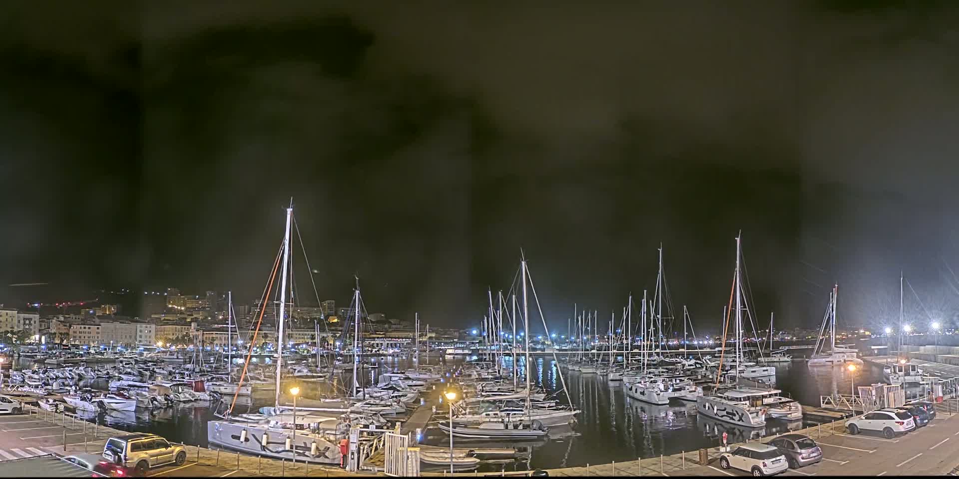 A marina filled with numerous sailboats and motorboats is illuminated at night under a dark, cloudy sky.