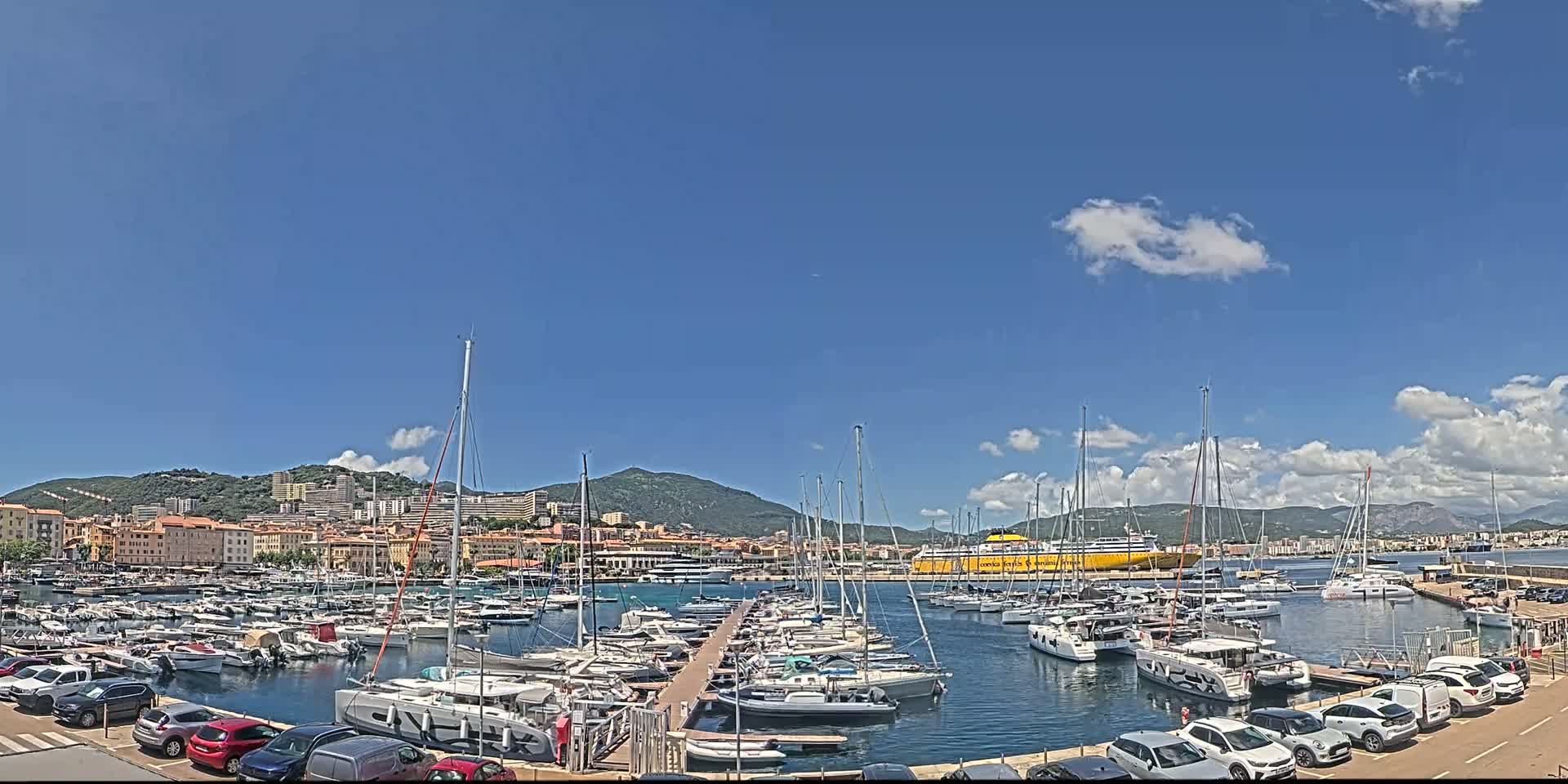 A marina filled with sailboats and motorboats is situated in front of a town nestled against a mountain range under a mostly sunny sky with a few scattered clouds.