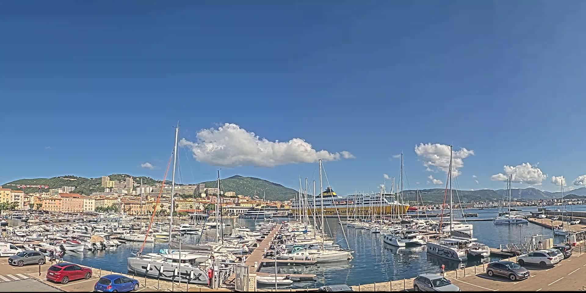 A marina filled with sailboats and yachts is nestled against a city backdrop under a mostly sunny sky with a few fluffy clouds.