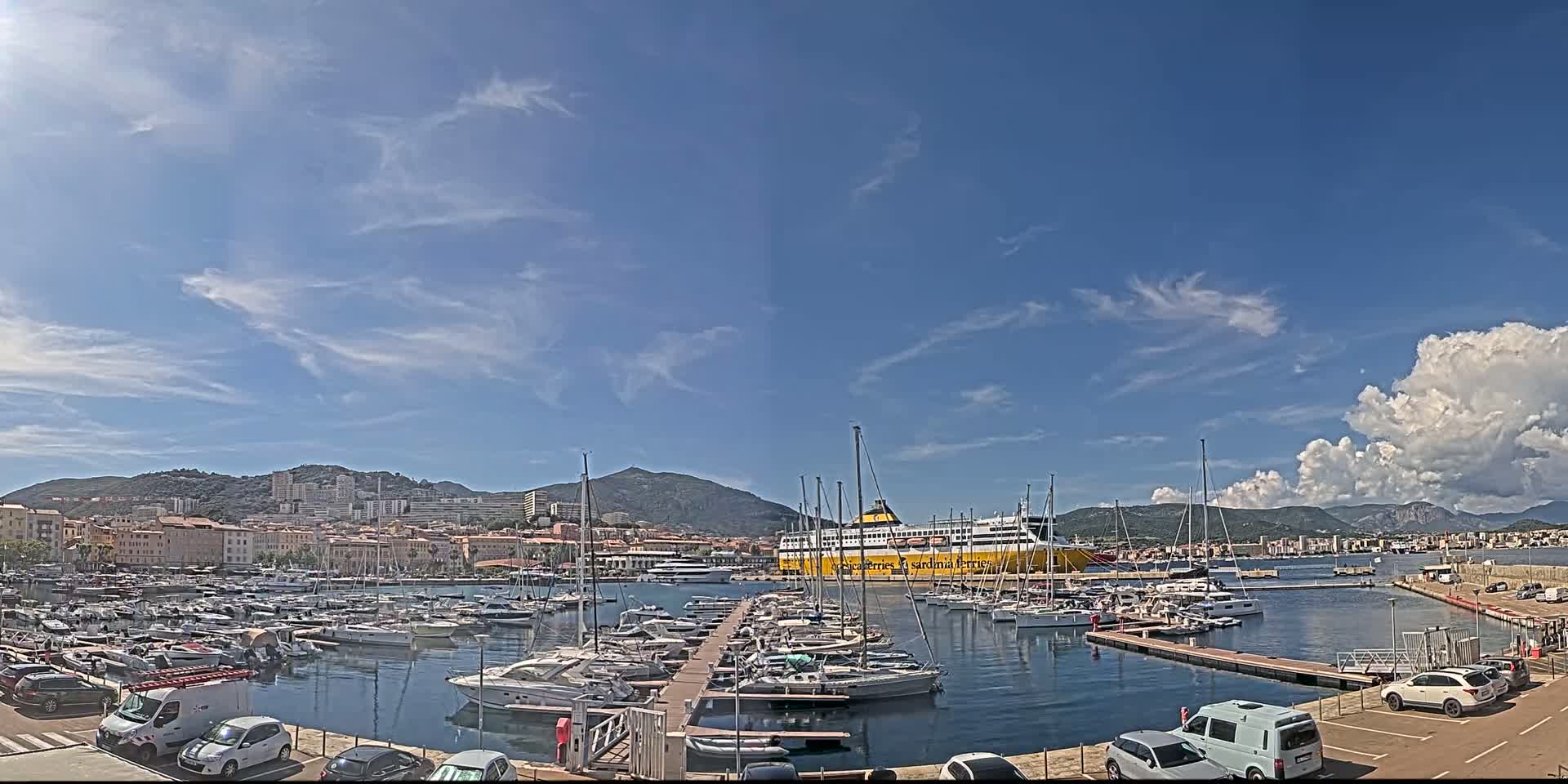 A marina filled with numerous yachts and sailboats is situated near a city nestled against mountains under a mostly sunny sky with scattered clouds.