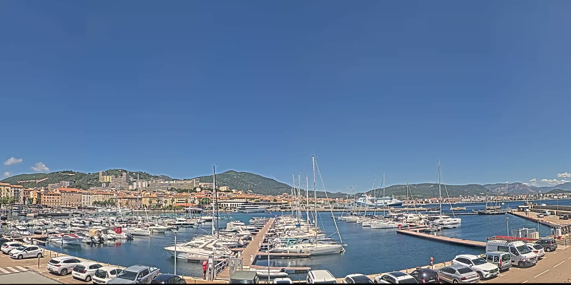A marina filled with boats is situated in front of a city, backed by hills under a clear blue sky.