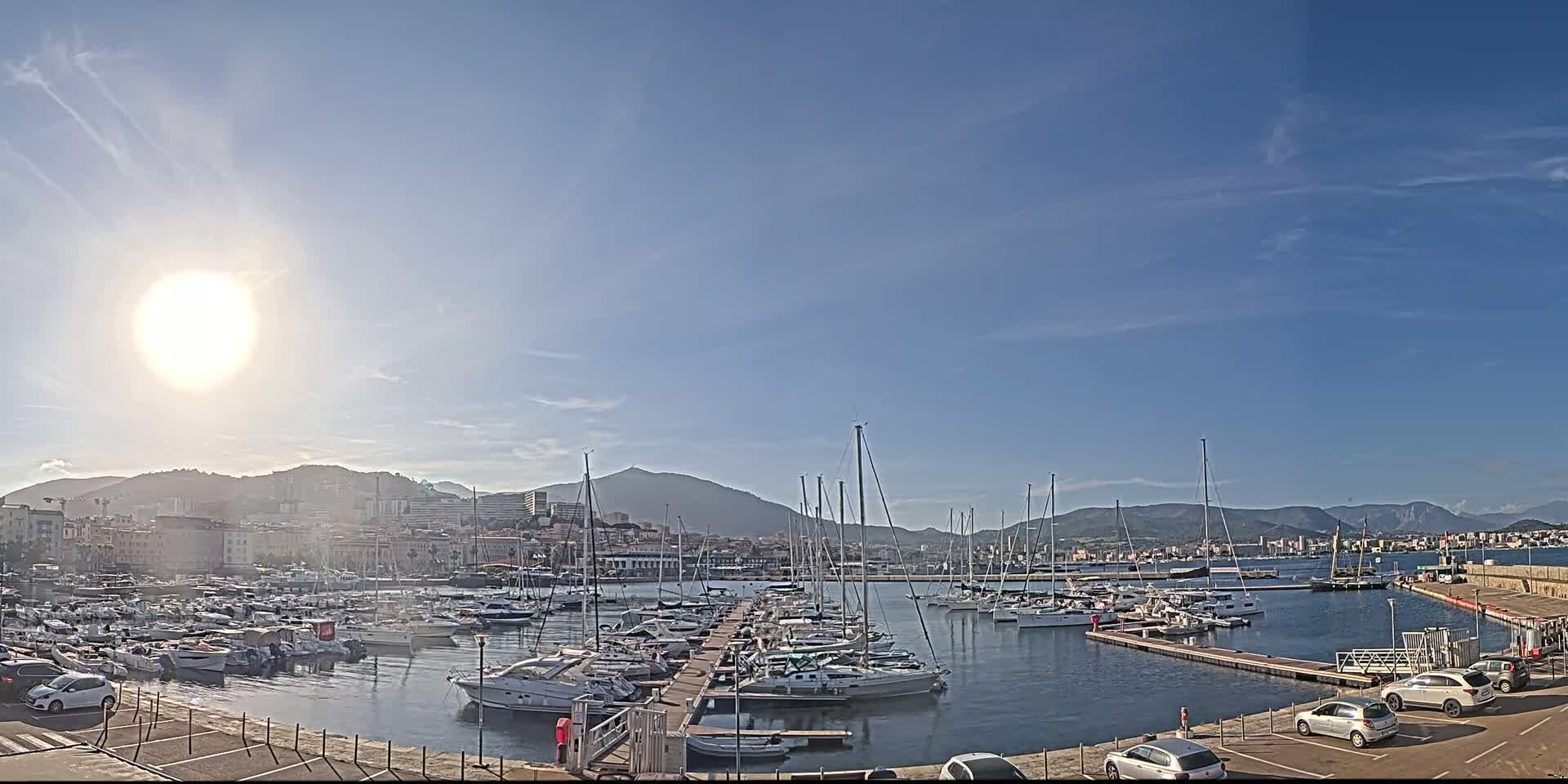 A sunny day reveals a harbor filled with numerous boats and yachts, docked alongside a city nestled against a mountain range.